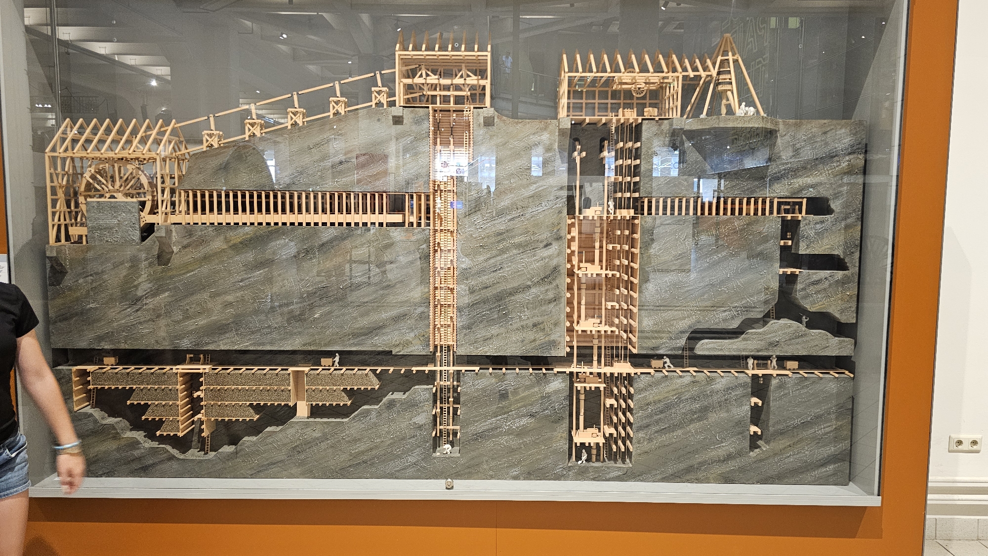 This is a detailed diorama of a cross-section of a mining operation, likely a historical exhibit. The diorama depicts multiple levels of underground tunnels and structures constructed from wood, simulating mine shafts, support beams, and scaffolding. The rock face is textured to resemble natural stone. Small figures representing miners are placed throughout the diorama, engaged in various tasks such as working in tunnels, operating machinery, and moving materials. There are also wooden carts and equipment visible. In the upper portion of the diorama, there are visible structures resembling mine heads and a large wooden wheel, possibly part of a hoisting mechanism. The diorama is encased in glass and displayed in what appears to be a museum or exhibition hall, with an orange frame around the display and a white wall with electrical outlets in the background. The location is listed as Unknown, Unknown.