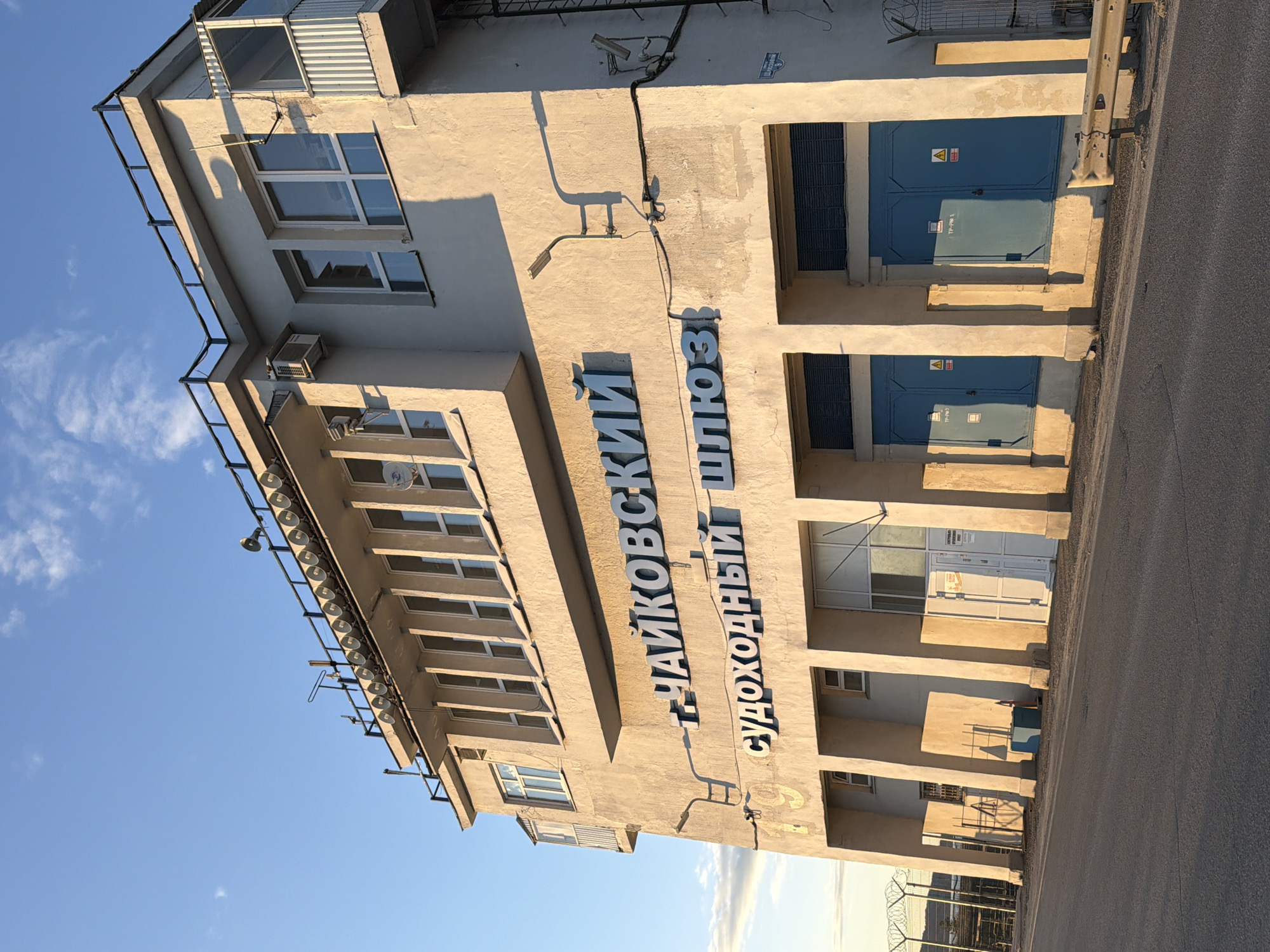 The image depicts a beige building in Chaikovsky, Russia. The building has multiple stories with rows of windows and a sign in Russian that reads "ЧАЙКОВСКИЙ СУДОХОДНЫЙ ШЛЮЗ" (Chaikovsky Shipping Lock). The building has blue electrical cabinets on its side. A paved road runs alongside the building. The sky is blue with light clouds. Barbed wire fencing is present in the foreground.