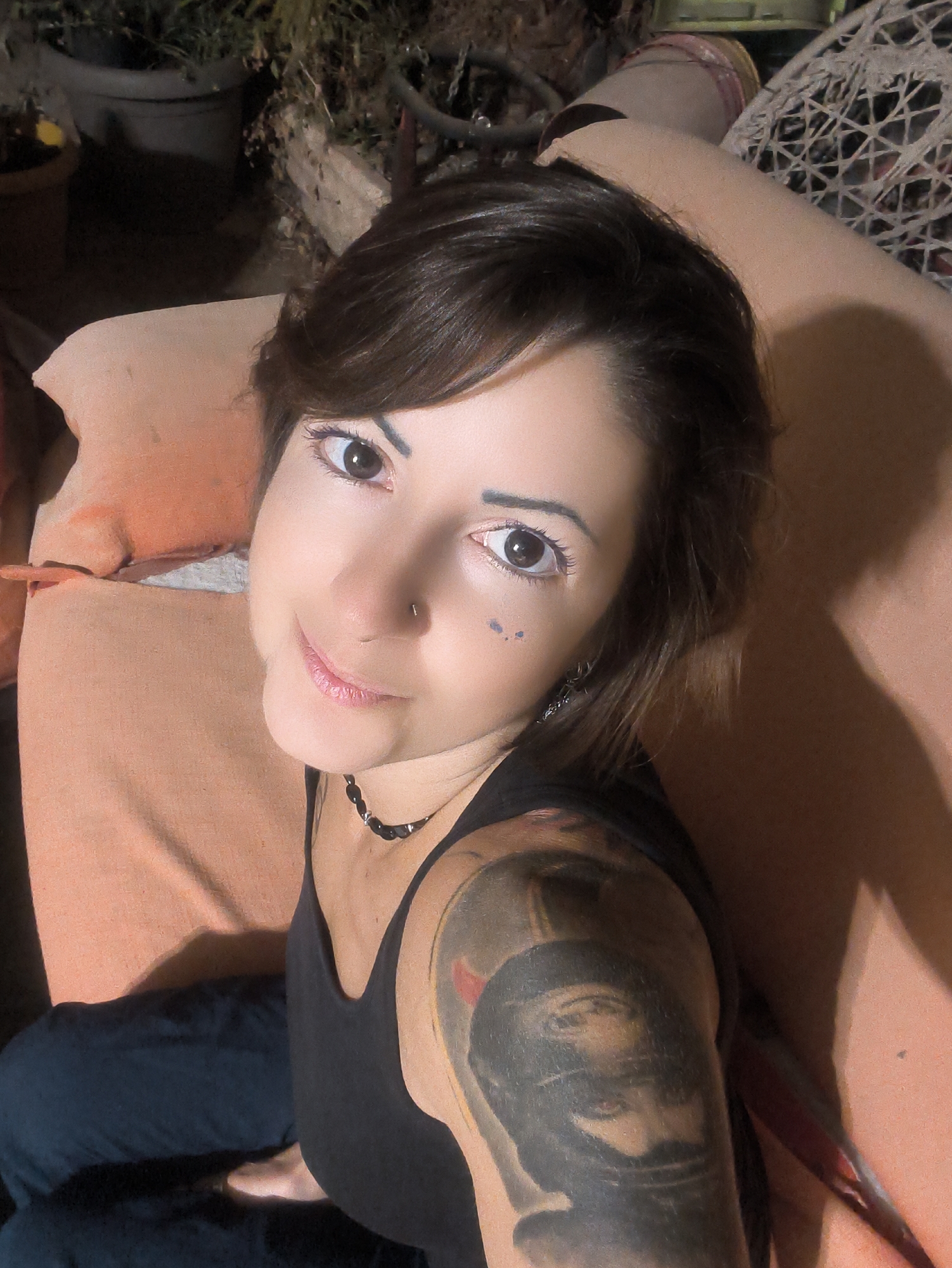The image shows a close-up, overhead shot of a woman lying down outdoors. She has brown hair, dark eyes, and is wearing makeup. She is wearing a black tank top and has a large tattoo on her upper left arm. She is also wearing a dark beaded necklace and has a nose ring. The background appears to be a garden with potted plants and outdoor furniture, suggesting a residential setting within the Lachish Regional Council, Israel.