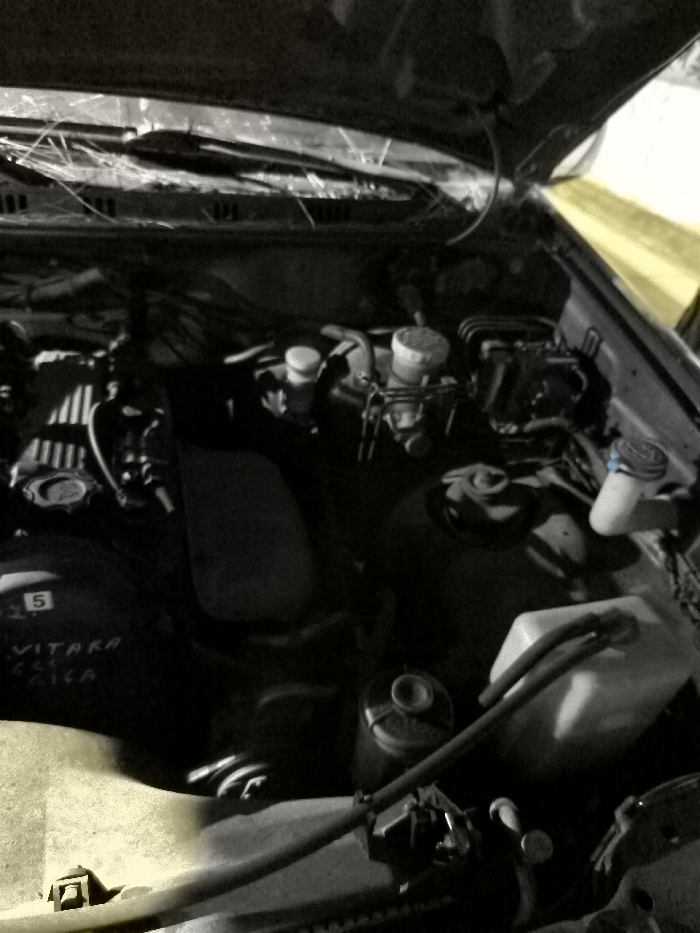 The media file displays an overhead view of an open car engine bay, revealing various components and mechanical parts. The main subjects are the vehicle's engine and associated systems, including fluid reservoirs (such as the brake fluid and coolant reservoirs), hoses, wiring, and the engine block. The underside of the car's hood is also visible, propped open.
The activity taking place is implied to be either an inspection, maintenance, or simply the car being parked with its hood open; no individuals are visible performing work.
Notable details include the lighting, which is generally dim, suggesting low-light conditions such as nighttime or an indoor garage with limited illumination. Yellow highlights are visible on the right side of the engine bay and on the bottom left, indicating an artificial light source, possibly a flashlight, illuminating specific areas. Debris, resembling dry leaves or twigs, is scattered across the windshield cowl area, suggesting the car may have been exposed to outdoor elements.
Visible text on the engine block or a component near it clearly identifies the vehicle as a "VITARA CLASSICA," with the number "5" also present above this text. One of the fluid reservoir caps on the upper right has a blue color or blue markings. There are no visual cues within the image itself to specifically indicate the provided location context of Iraklio, Greece. The overall mood is utilitarian and focused on the mechanical aspects of the vehicle.