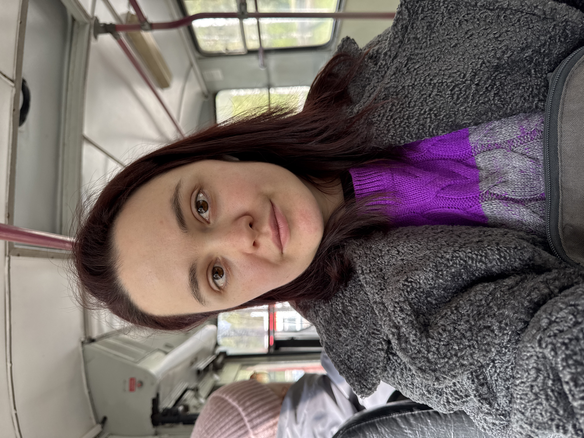 A woman with brown hair is on a bus. She is wearing a gray fleece jacket over a purple and gray sweater. The bus is passing through a city. The city might be Izhevsk, Russia, as requested.