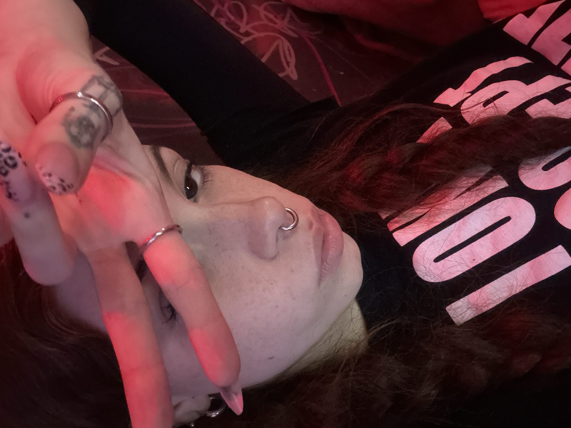 A young woman is lying down, possibly on a bed, as suggested by the bedding visible in the background. She's wearing a black top with large, light-colored letters printed on it. Her brown hair is spread around her head. She has a nose ring and is wearing rings on her fingers. Her left hand is raised, partially obscuring her face. The image appears to be taken in an indoor setting with somewhat subdued lighting, possibly with a red light source affecting the color cast. There is no information within the image to suggest a location within the city of Buenos Aires, Argentina.