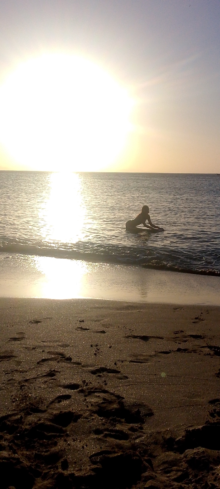 A figure kneels in shallow water at the beach, silhouetted against a bright, hazy sunset. The sun reflects intensely on the water's surface, creating a shimmering effect. The calm sea stretches to the horizon under a light-colored sky. The foreground is dominated by sandy beach with numerous footprints imprinted on the surface. The image is overall warm and bright.
