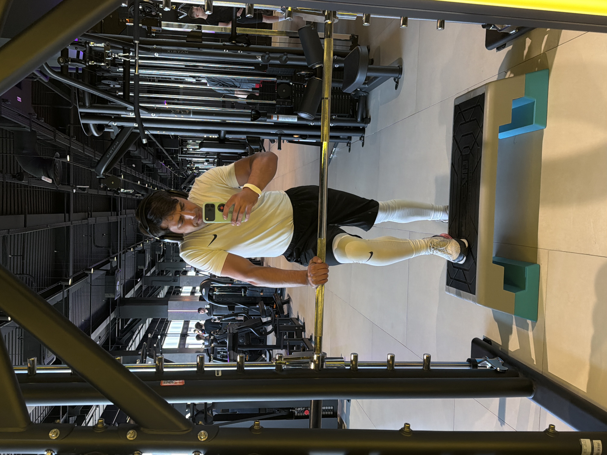 This image, originally rotated, displays a man posing for a photograph in a modern gymnasium. He stands upright in front of a piece of exercise equipment, possibly a Smith machine or power rack, holding a golden-colored barbell with both hands at waist height. His gaze is directed downwards at a light green smartphone he holds in his left hand, suggesting he is capturing a selfie or reviewing the photo.
The man is dressed in athletic wear, including a light-colored (likely white or cream) t-shirt featuring a black Nike swoosh logo on the left chest. He wears black shorts, white athletic compression sleeves or long socks covering his calves, and athletic shoes. A white wristband is visible on his right wrist, and he has black over-ear headphones.
The background reveals a spacious and well-lit gym interior. Numerous black and metallic gym machines are visible, some reflected in mirrored surfaces. The ceiling features dark, industrial-style beams and structures. The floor is a light, neutral color, possibly concrete or tiled. The reflections also show other individuals in the gym. The word "MATRIX" is visible in reverse text on some of the equipment, indicating the brand. The Nike logo is also present on his leg wear. The scene suggests a contemporary fitness facility in Krasnoyarsk, Russia.