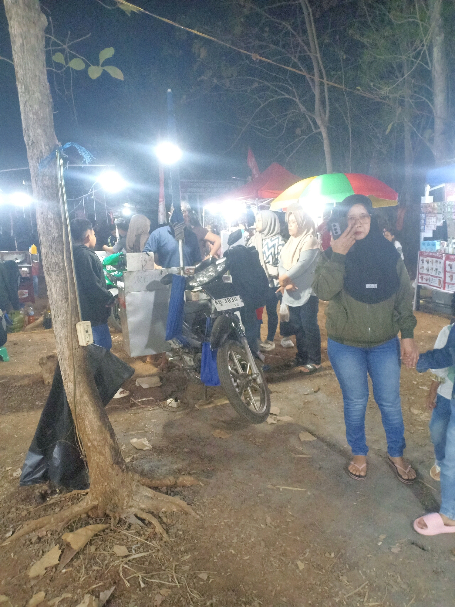 This is a nighttime, outdoor scene in what appears to be a street market or food stall area in Tasikhargo, Indonesia. A woman wearing a dark hijab and a green jacket is in the foreground, holding up her left hand with two fingers extended, possibly posing for a photo. She is wearing blue jeans and flip-flops. Behind her, a motorcycle with license plate "AB 3836 K" is parked. Several other people, mostly women wearing hijabs, are visible in the mid-ground, some standing and some interacting near food stalls. Colorful umbrellas are also present. The ground is dirt and debris, and trees with visible roots are in the left and background. Overhead lights illuminate the scene, creating a lively but somewhat hazy atmosphere.