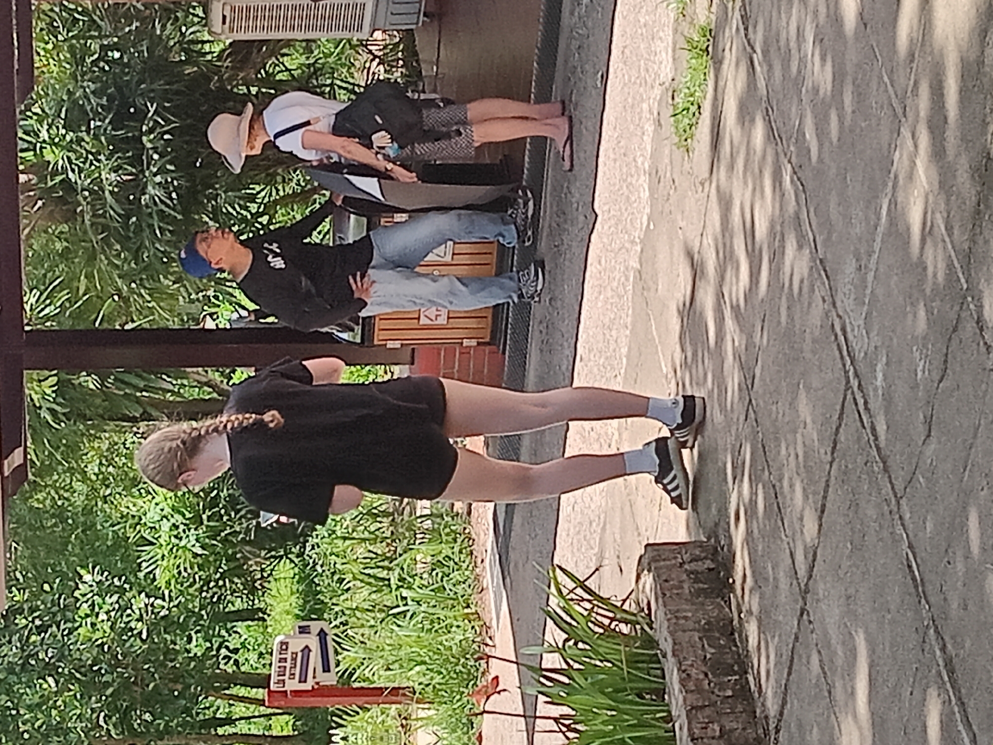 The image depicts an outdoor scene at the Marble Mountains in Da Nang, Vietnam. In the foreground, a young person with blonde hair in a braid, wearing a black t-shirt and black shorts, stands on a paved walkway. They are facing away from the camera, with their back to a brick planter box filled with greenery. To the right, two adults are engaged in conversation. A person wearing a blue baseball cap and a black long-sleeved shirt is gesturing, while a person wearing a straw hat and a white shirt faces them. Behind them, a sign with Vietnamese text and arrows indicates "ENTRANCE." The environment is characterized by lush green foliage, suggesting a tropical or subtropical climate. The ground is paved with concrete tiles, and there is dappled sunlight casting shadows from the trees overhead. A metal railing and part of a building or structure are visible in the background. The overall scene suggests a tourist location with people exploring the area.