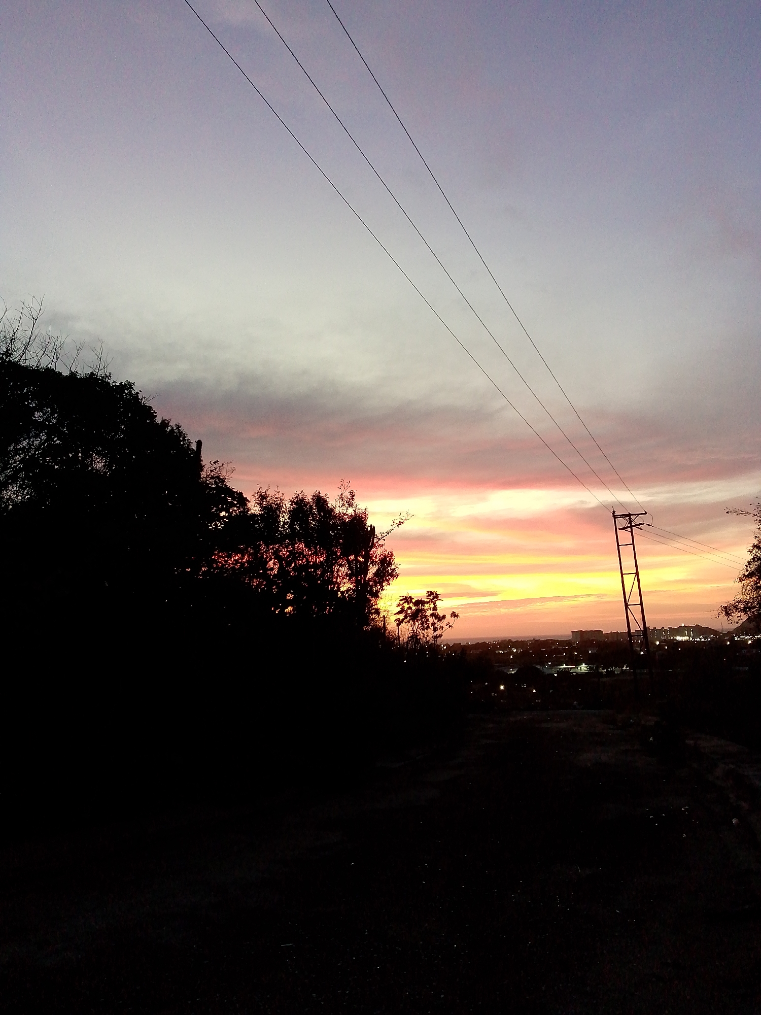 The image depicts a sunset scene in Altagracia, Venezuela. The foreground is dominated by dark silhouettes of trees and a dirt road. In the middle ground, there's a power pole with electrical lines extending across the sky. The background features a cityscape illuminated by streetlights and buildings, silhouetted against the colorful sunset. The sky exhibits a gradient of colors, ranging from yellow and orange near the horizon to pink and purple higher up. No people are visible in the image.