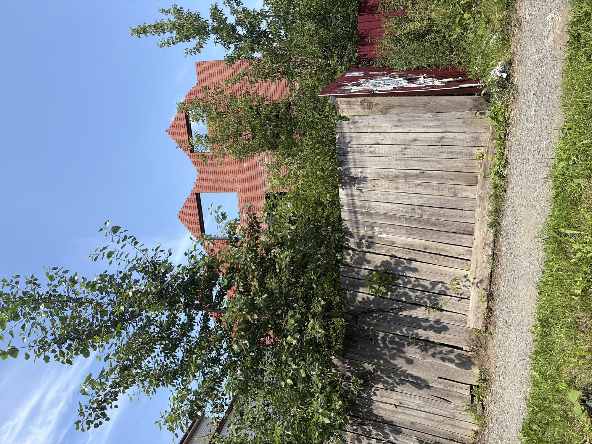 The image depicts an unfinished brick building under a clear blue sky. A wooden fence runs across the foreground, with a section of red metal fencing to the right. Trees and green foliage are abundant, obscuring parts of the building. The ground in front of the fence is a gravel path, bordered by green grass. This scene is located in the city of Aramil, Russia.