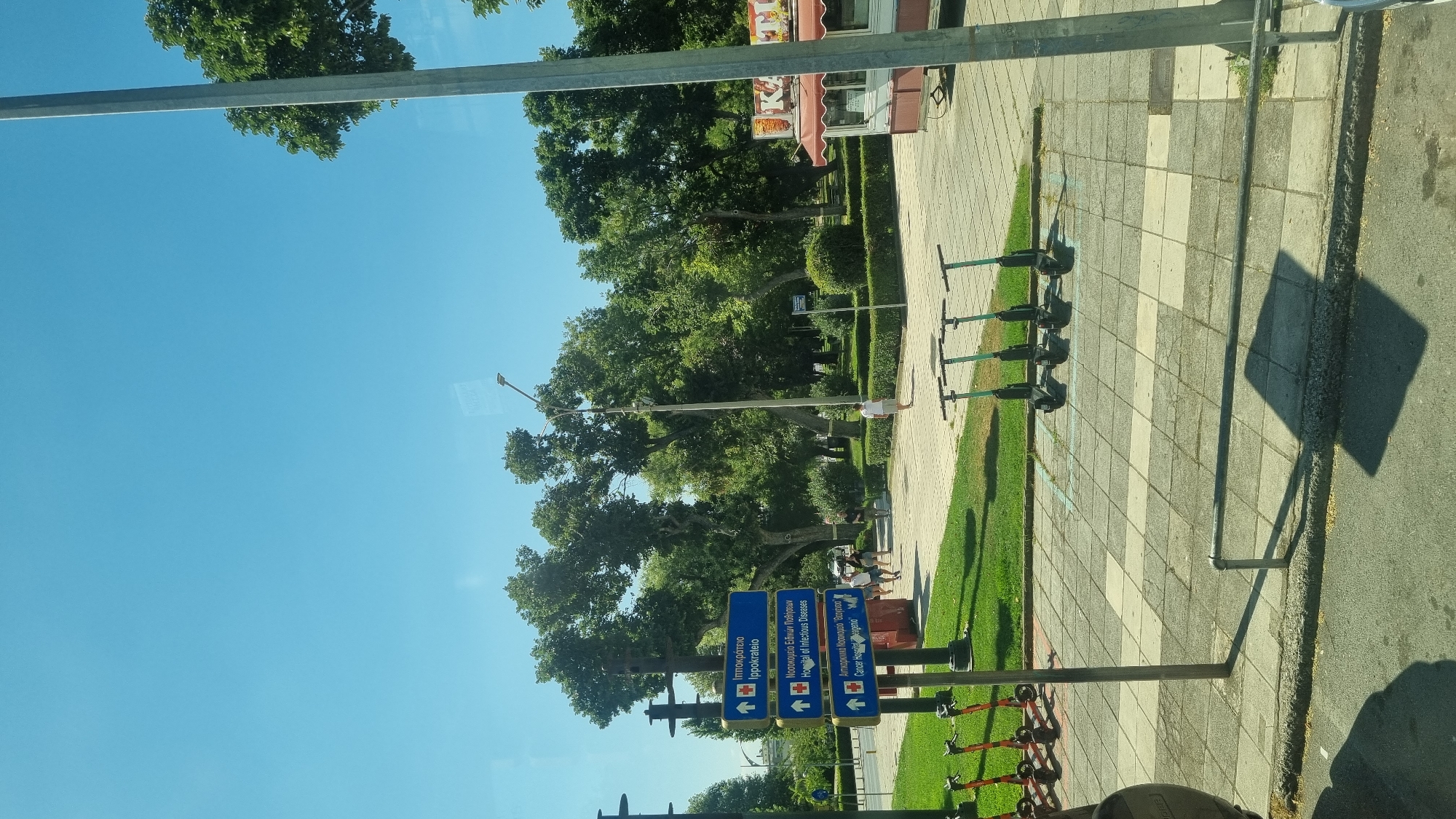 The image is taken from a moving vehicle, likely a bus, looking out a window. The scene is an outdoor urban environment with a clear blue sky. In the foreground, there are several electric scooters parked on a tiled sidewalk. Behind the scooters, a patch of green grass is visible, with a pathway leading further into a park.
Dominating the middle ground are several tall trees with lush green foliage. A street signpost is prominent on the left, displaying directional arrows and text in Greek and English indicating "Hippokrateio / Hippocrates", "Hospital of Infectious Diseases", and "Cancer Hospital". People are walking on the pathway in the background. To the right, a small food cart with a red awning is visible. The overall setting appears to be a city park in Thessaloniki Municipal Unit, Greece, possibly near a hospital complex given the signage.