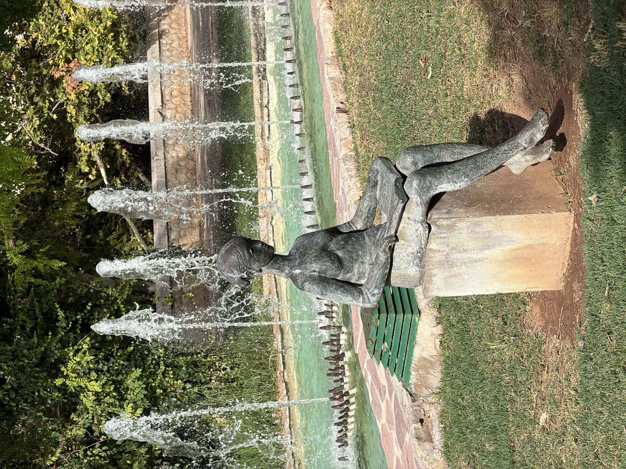 A bronze statue of a young woman reading a book sits on a concrete pedestal in a park. The statue is in the foreground, with a fountain and lush greenery in the background. The fountain has multiple streams of water spraying upwards, creating a misty effect. The woman in the statue is depicted in a seated position, with her legs crossed and her arms holding an open book. Her hair is styled in a bun, and her gaze is directed downwards at the book. The statue appears to be made of weathered bronze, with a patina that gives it a greenish hue. The concrete pedestal is rough and weathered, with some dirt and grass growing around its base. The park environment is well-maintained, with manicured grass and mature trees providing shade. The overall scene evokes a sense of tranquility and intellectual pursuit.