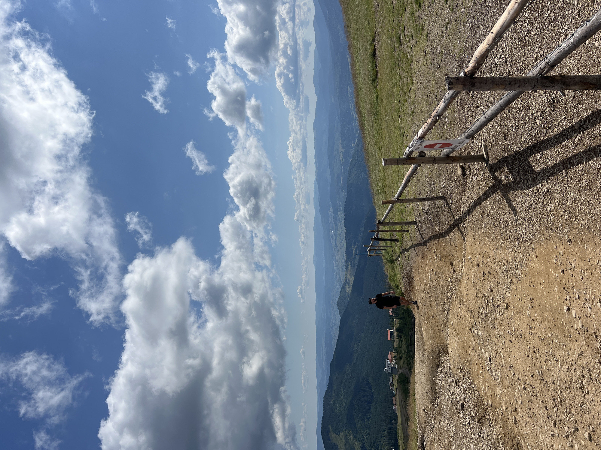 A man wearing a black t-shirt and shorts walks down a dirt path on a grassy hillside. He is walking away from the viewer. The path is lined with a rustic wooden fence on the right side, with a red and white circular sign attached to it. In the background, there are rolling hills covered in green trees, with buildings visible on the lower slopes. In the far distance, more mountain ranges are visible under a partly cloudy blue sky. The image appears to be taken in Chorna Tysa, Ukraine, given the mountainous terrain and architecture.