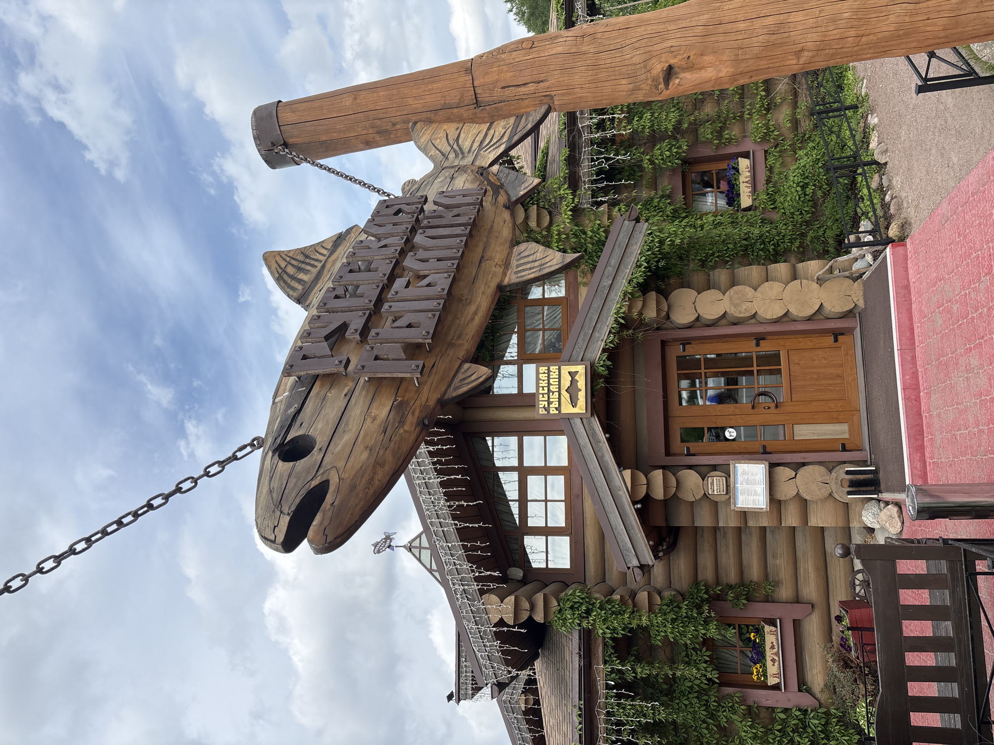 The image captures the exterior of a rustic, log cabin-style building, likely a restaurant or lodge, under a cloudy daytime sky in Saint Petersburg, Russia. A large, carved wooden fish serves as a prominent sign, suspended by heavy metal chains from a robust wooden beam. The text inscribed on the fish's body, and reiterated on a smaller sign below it, clearly spells out "РУССКАЯ РЫБАЛКА" (RUSSKAYA RYBALKA), translating to "Russian Fishing" or "Russian Fishery."
The building's facade is constructed from horizontal logs and is partially enveloped by lush green climbing plants, creating a natural and inviting appearance. Multiple windows with brown frames are visible, and decorative string lights are draped along the roofline, suggesting a cozy ambiance, particularly for evening hours. Near the main entrance, a smaller, rectangular sign with indistinct text is present, possibly displaying a menu or operational details. The ground in front of the establishment consists of a mix of gravel or dirt with some interspersed rocks, and a red paved or brick area is visible to the right. No people are present in the scene. The overall impression is that of a charming, themed establishment with a strong traditional Russian aesthetic.