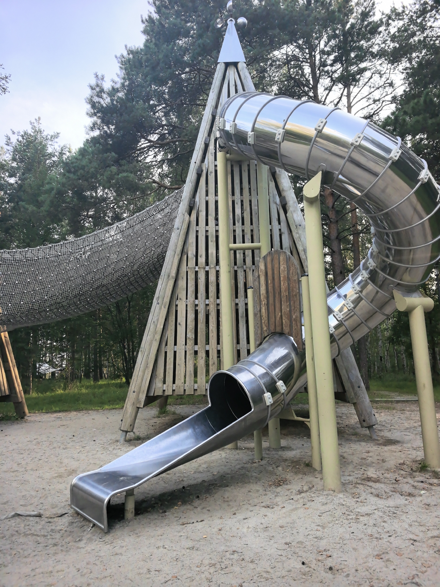 The image shows a modern playground structure made of wood and metal, located in an outdoor setting with trees and sand. The structure features a tall, pyramid-shaped wooden tower with vertical planks, topped with a metallic cone and spheres. A large, curved, tubular metal slide descends from the upper part of the tower. In front of this, a shorter, straight metal slide extends to the ground. A net tunnel is attached to the side of the wooden tower, leading away to the left. The ground is covered with sand and some fallen leaves. The scene is bathed in natural light, suggesting it is daytime. The playground appears to be in a forested area, possibly a park or a recreational space. The specific location within Verkhnyaya Pyshma, Russia, is not visually discernible from the image alone, but it depicts a public recreational facility. There are no people visible in the image.