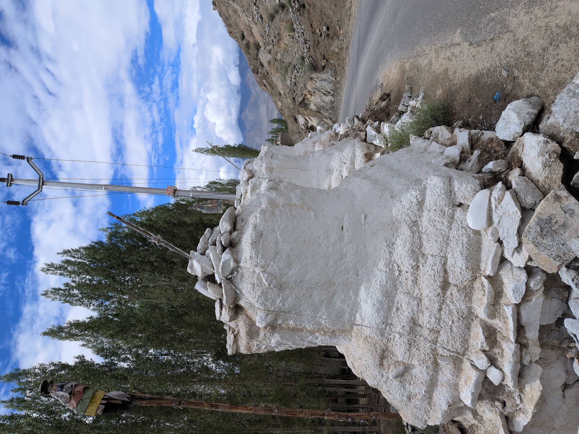 The image shows a roadside scene in Leh, India, on a cloudy day. A rough, white-plastered stone structure stands in the foreground, decorated with loose white stones at its base and top. To the left, a wooden pole with prayer flags is visible, with a line of tall green trees behind it. A utility pole with electrical wires runs vertically behind the white structure. A paved road curves to the right, bordered by a rocky, barren hillside. The background features distant mountains partially obscured by clouds. The scene depicts a blend of man-made structures and natural landscape typical of the region.