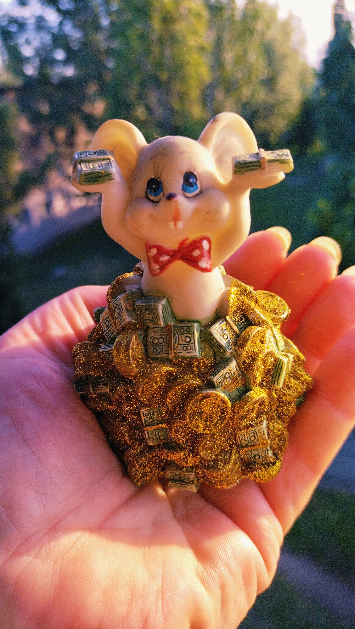 The image shows a close-up of a person's open palm holding a figurine of a mouse. The mouse is light-colored with blue eyes and a red bow tie with white polka dots. It is adorned with miniature stacks of what appear to be US dollar bills and gold coins, all covered in gold glitter. The mouse's ears also have stacks of bills on them. The background is softly blurred, showing green foliage, suggesting an outdoor setting, possibly a park or garden. The lighting suggests it is daytime, with sunlight casting a warm glow on the hand and figurine. There is no discernible indication of the location being in Horishni Plavni, Ukraine.