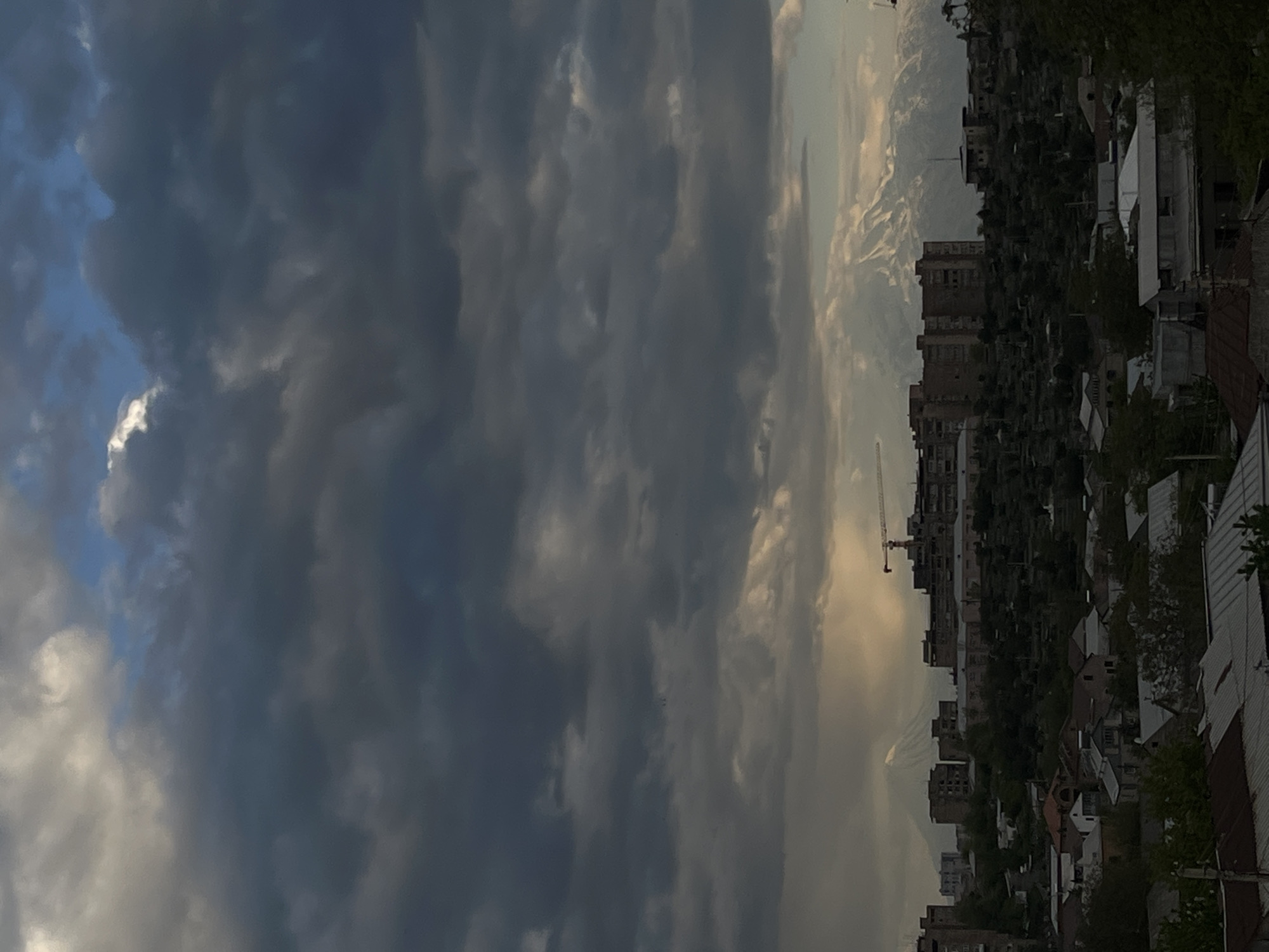 The image depicts an urban landscape under a dramatic sky. Heavy, dark storm clouds dominate the left side of the frame, contrasting with a lighter, brighter area on the right, suggesting the setting or rising of the sun.
Below the sky, there is a view of a town. Buildings of various sizes are visible, interspersed with trees and greenery. There appears to be a construction crane in the scene, indicating ongoing development.
In the distance, there are hints of mountains. The city of Arinj, Armenia has views of Mt. Ararat, so it is likely that those are the mountains in the background.
