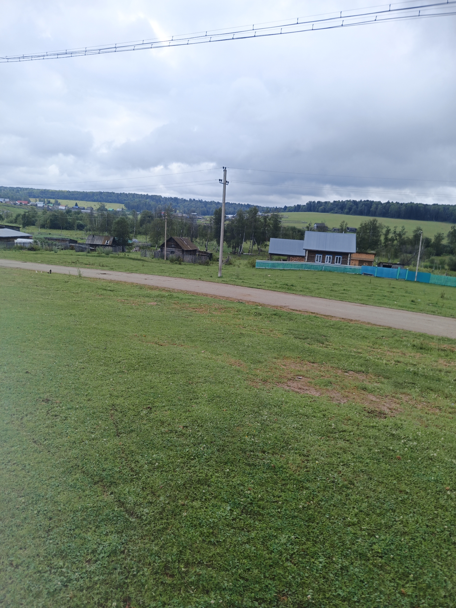 A pastoral scene unfolds in Islambaevo, Russia, under a cloudy sky. A dirt road cuts diagonally across the frame, bordered by a lush green lawn on the left and a more uneven, patchy lawn on the right. In the mid-ground, several wooden houses with weathered roofs are scattered amongst trees and fences. One house stands out with a corrugated metal roof and several windows. A vibrant teal fence encloses a portion of the property. In the distance, rolling hills are covered with a dense forest. The image captures a quiet, rural moment, devoid of human activity.