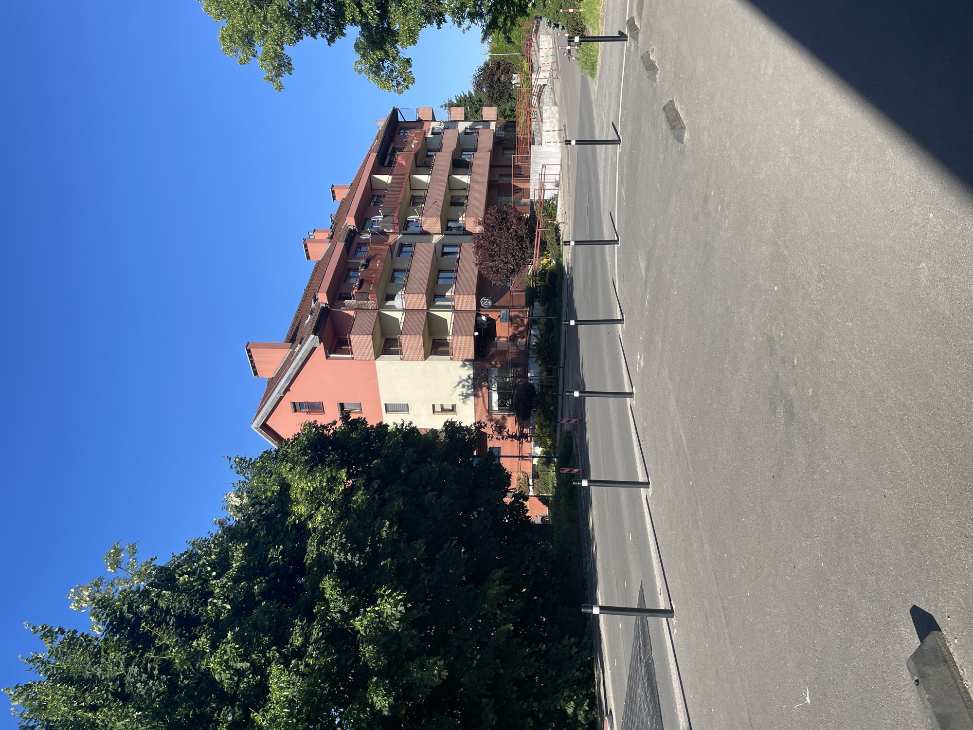 A medium-angle view reveals a six-story building in Wrocław, Poland. The building features a mix of peach and off-white facade sections, with brown brick balconies on each floor. Several satellite dishes are attached to the building's exterior. A row of evenly spaced, dark bollards lines the street in front of the building, separating it from a paved area. Trees with lush green foliage frame the lower left portion of the image, while the clear blue sky dominates the upper left. A shadow obscures the top right corner of the paved area. The composition focuses on the architecture and urban landscape.