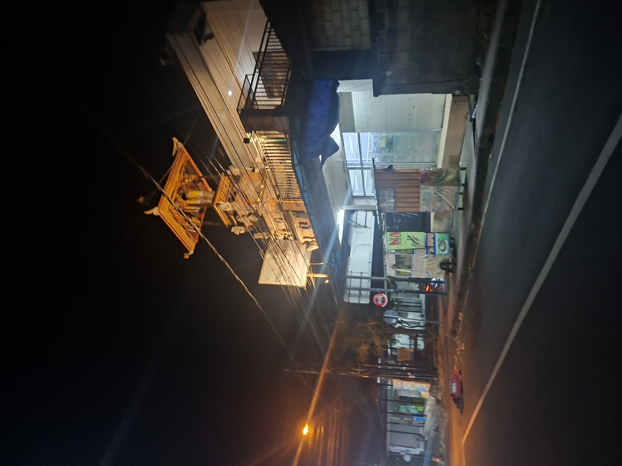 The image shows a street scene at night in Blahbatuh, Indonesia. A street light illuminates the asphalt road and the side of a building. The building appears to be a shop or a small business, with goods displayed in windows and on shelves. There are signs with text in what appears to be Indonesian. In the foreground, a yellow and orange structure, possibly an altar or a decorative piece, hangs from wires. The overall atmosphere is that of a busy street at night, with lights and activity. The image is slightly tilted, giving a dynamic perspective.