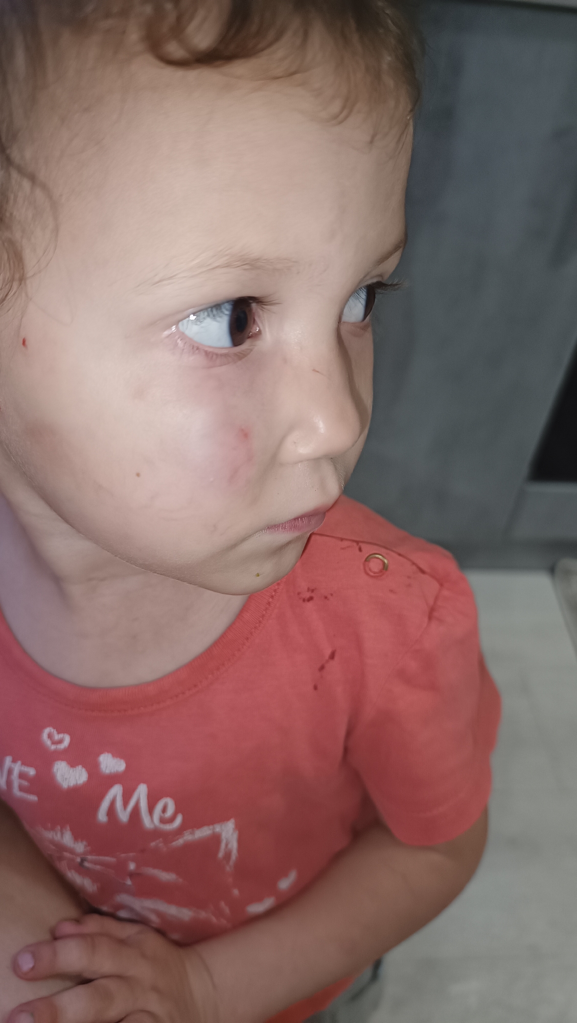 A close-up, profile shot shows a young Caucasian child looking to the right. The child has light skin, brown eyes, and curly brown hair. The child is wearing a coral-colored t-shirt with white graphic print. The t-shirt appears to have some reddish-brown stains on it, as does the child's face and neck. The background is out of focus and appears to be an indoor setting with a gray wall and possibly a countertop. The image does not provide any information about the city of Chaikovsky, Russia.