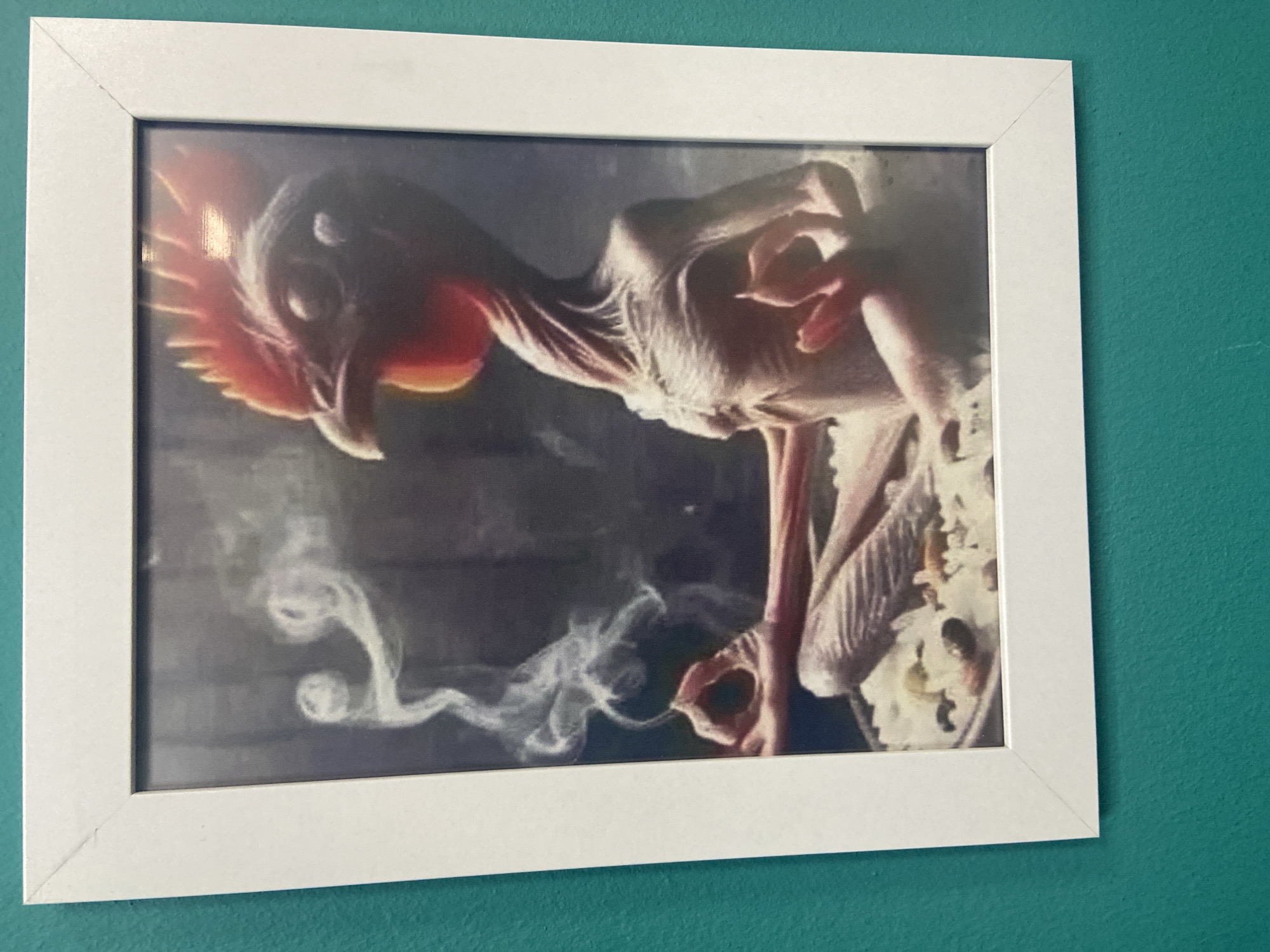 This is a photograph of a framed picture hanging on a teal-colored wall. The framed picture appears to be a print or painting depicting a mythical or fantastical creature. The creature is thin and pale, with a long neck and a prominent, bird-like head that has red, feather-like protrusions around it. It has slender limbs and delicate, elongated fingers. In the foreground, wisps of white smoke or mist are swirling, creating a dramatic and atmospheric effect. The background of the artwork is dark and indistinct, focusing attention on the creature and the smoke. The frame is simple and white, with clean lines. The teal wall provides a vibrant contrast to the white frame and the imagery within. There is no visible text in the image. The scene depicted in the artwork is one of mystery and fantasy, with no clear indication of time of day, weather, or a specific location beyond the artwork itself.