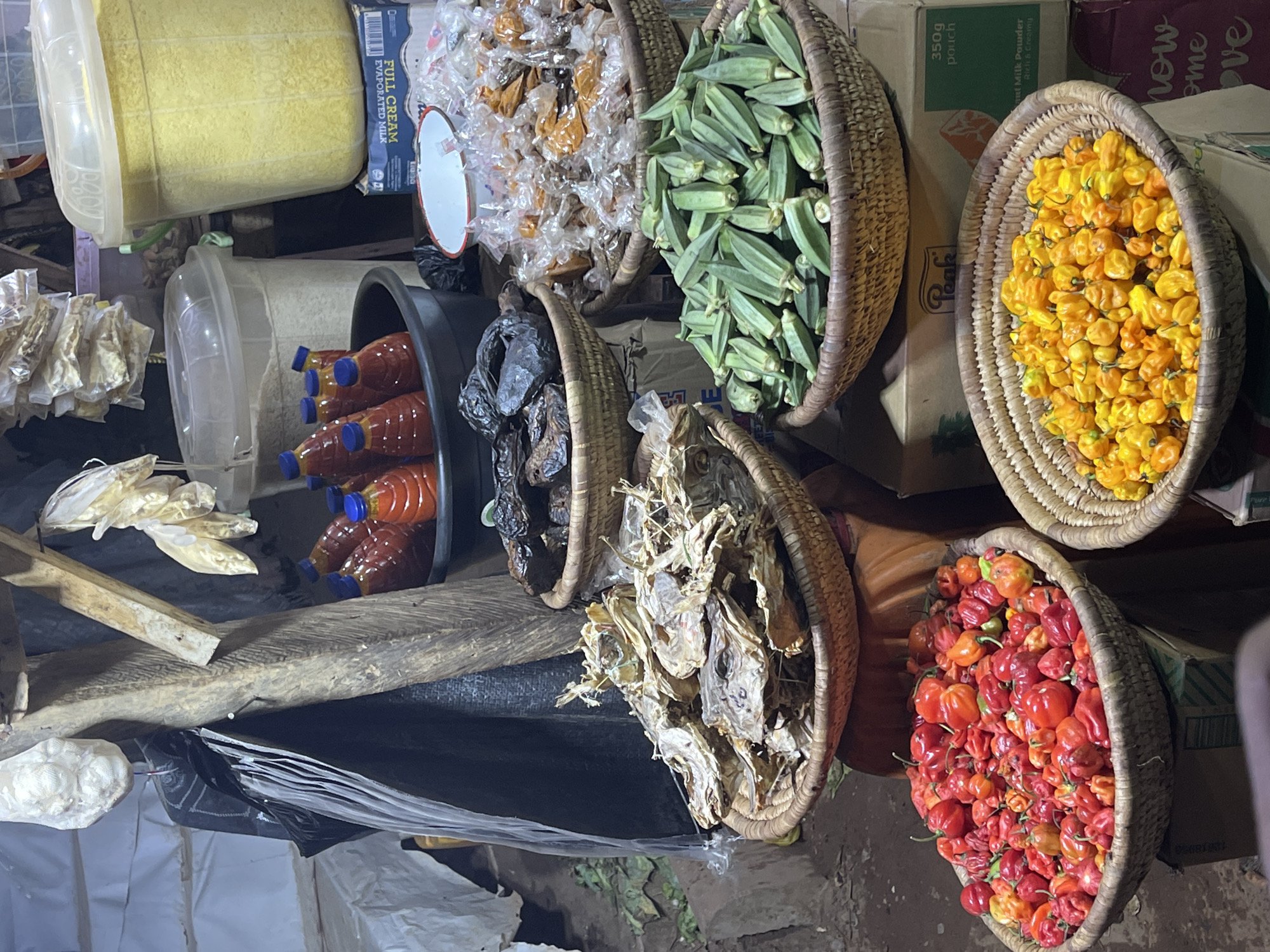 This bustling market scene in Abuja, Nigeria, showcases a variety of food items meticulously arranged for sale. The image is captured from an elevated perspective, looking down into several woven baskets and containers brimming with fresh produce and dried goods.
Prominent subjects include a large quantity of fresh green okra in one basket, vibrant yellow habanero peppers in another, and an abundant display of red habanero peppers in a third. Other baskets hold dried fish or similar preserved seafood, and individually wrapped portions of what appear to be dried snacks or spices.
Various containers are also visible: a large clear plastic bin filled with a yellow granular substance, likely garri or maize flour, and a black bucket holding multiple plastic bottles of red palm oil. Bags containing dried ingredients are stacked on shelves or surfaces. The setting suggests a busy, local food market, with goods piled high, some on cardboard boxes serving as makeshift stands.
Notable details include the bright, artificial lighting that illuminates the stall, indicating it might be an indoor market, evening, or night. The vibrant colours of the peppers and palm oil contrast with the earthy tones of the woven baskets and dried items.
Visible text includes "FULL CREAM EVAPORATED MILK," "Peak" (a brand name), "350g pouch," and "Instant Milk Powder Rich & Creamy" on packaging, along with a partial "now ome ove" which might be part of a larger phrase on a bag. The number "6080" is also visible. The absence of people suggests a focus on the goods themselves or was taken during a lull in activity.
