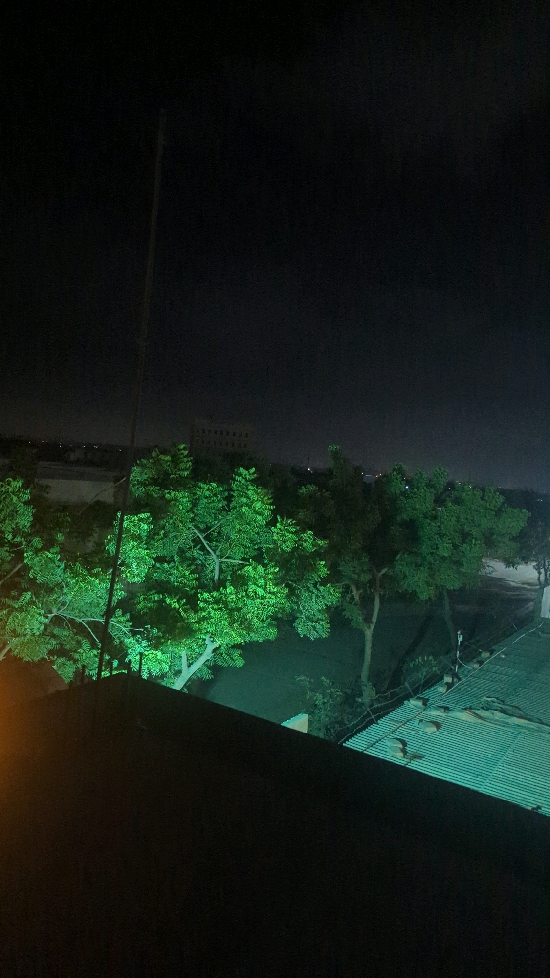 The image shows a night scene with trees illuminated by green light. In the foreground, there is a dark rooftop. Beyond the trees, a building is visible in the distance, and a corrugated metal roof is present to the right. The scene appears to be located in the city of Hub, Pakistan, based on the context provided. No people or specific actions are depicted. The environment is dark, suggesting it is nighttime.