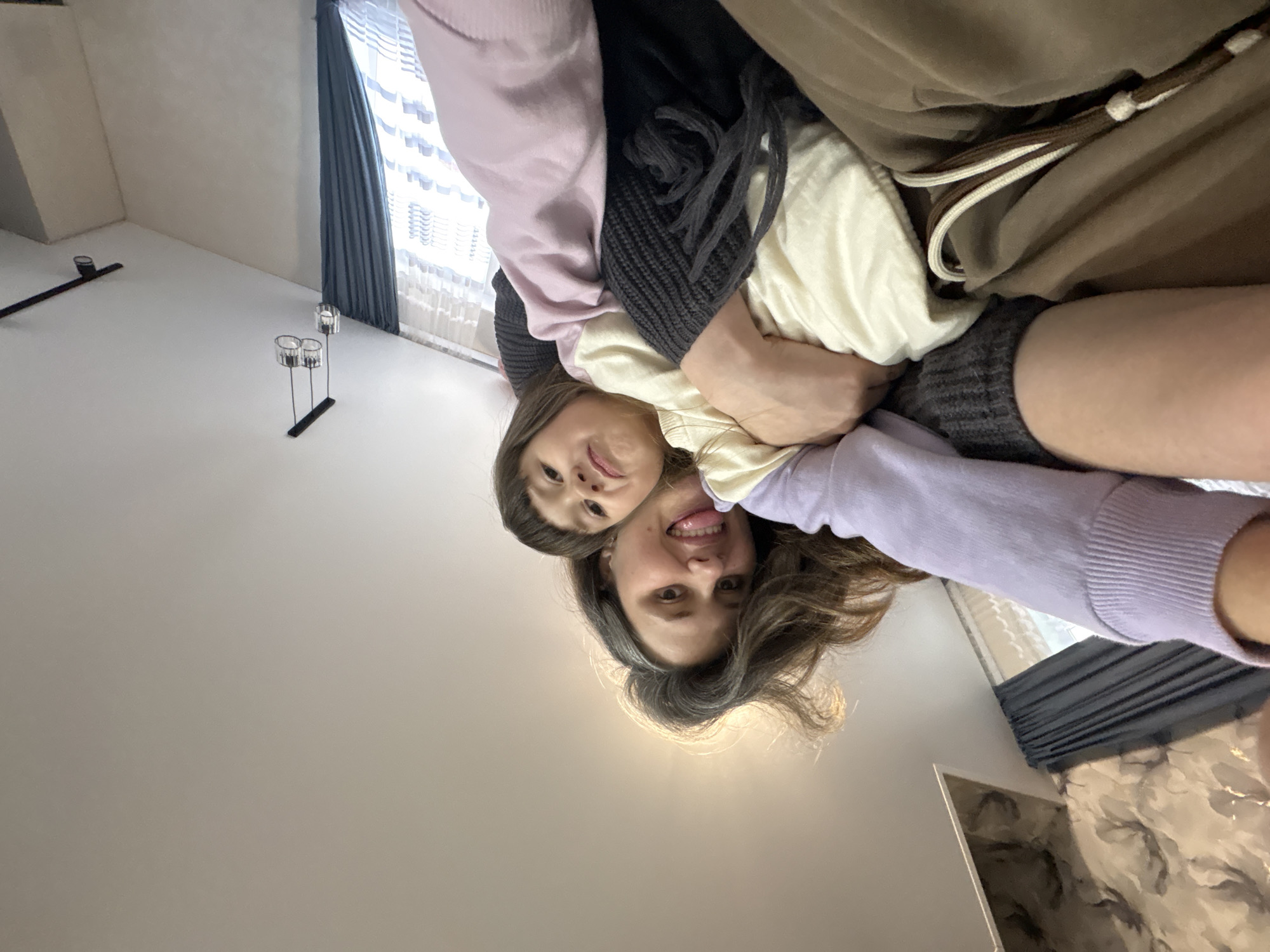 A photograph depicts two people, likely a mother and daughter, embracing closely from a low-angle perspective within an interior space. The younger person, possibly a child, is positioned slightly above the older individual. Both are facing towards the camera. A variety of clothing fabrics in pastel shades of pink and lilac, alongside darker tones of gray and beige, are obscuring much of their forms.

The setting appears to be inside a residential building with minimalist décor. In the background, the room is visible, displaying features like white walls, gray curtains, and a metal candelabra. Considering the latitude and architecture shown, it is plausible the location is Prikamskii, Russia. The action is a close embrace.