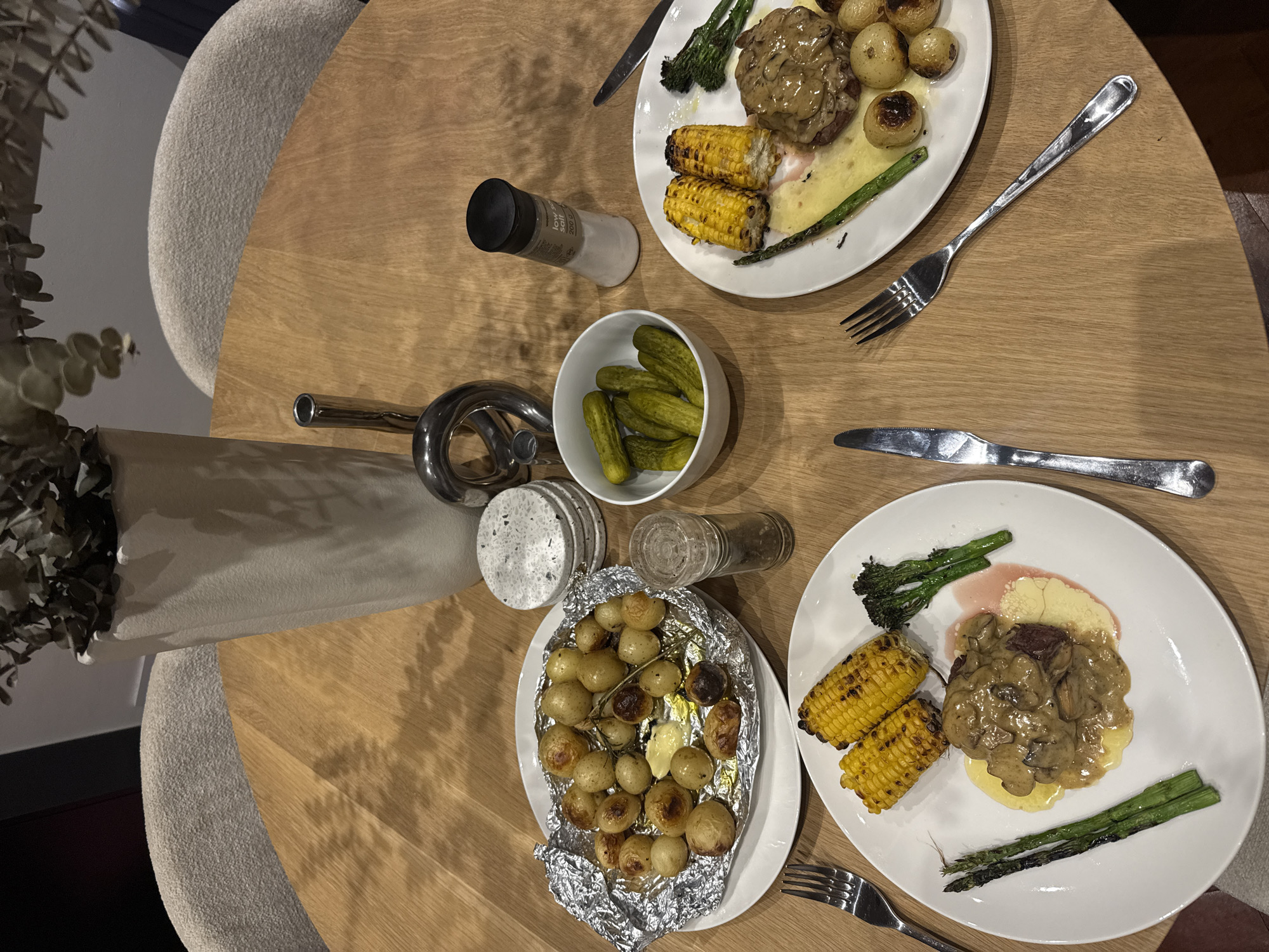 A high-angle, indoor shot depicts a wooden round table setting for two, possibly in Cape Town, South Africa, given the food styles. Each setting includes a white plate with a portion of steak covered in mushroom sauce, grilled corn on the cob, asparagus, and broccolini. One setting has roasted potatoes on foil. A small white bowl contains dill pickles. Salt and pepper shakers, along with cutlery (forks and knives), are present. A decorative metal object and a vase holding foliage sit on the table. The table is surrounded by upholstered chairs.