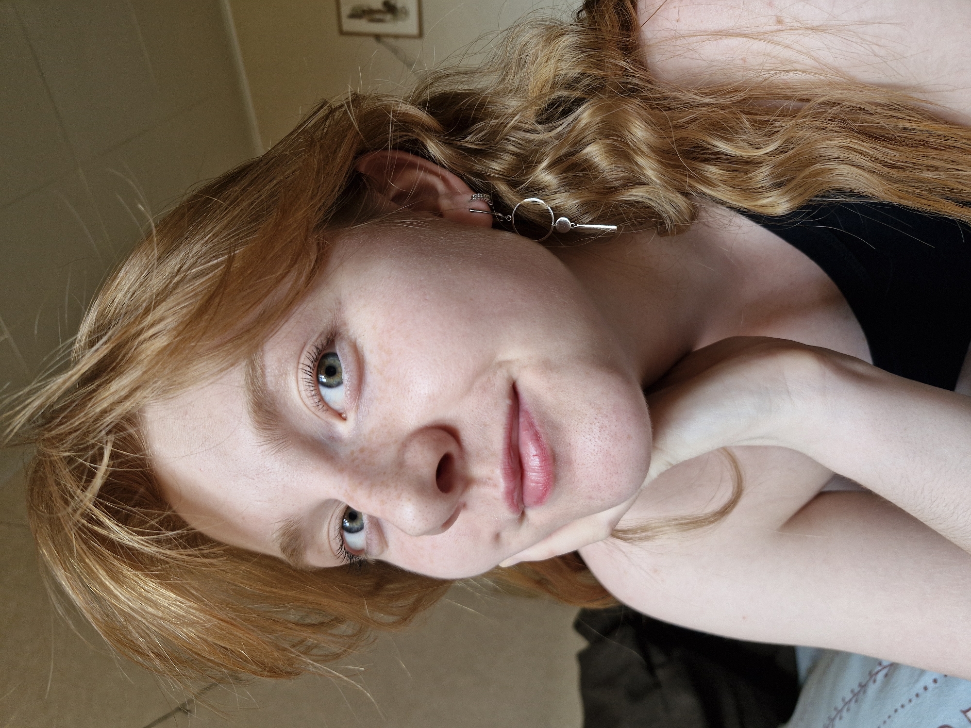 A close-up, rotated image shows a young, fair-skinned woman with auburn, curly hair and freckles. She has light green eyes and wears a modern geometric earring on her left ear. The woman is looking up towards the left of the frame with a slight smile. Her arms are crossed and supporting her head. She is wearing a black top. The background consists of a neutral-toned wall with a framed picture hanging on it. There is no explicit information provided in the image that identifies a location in Yekaterinburg, Russia.