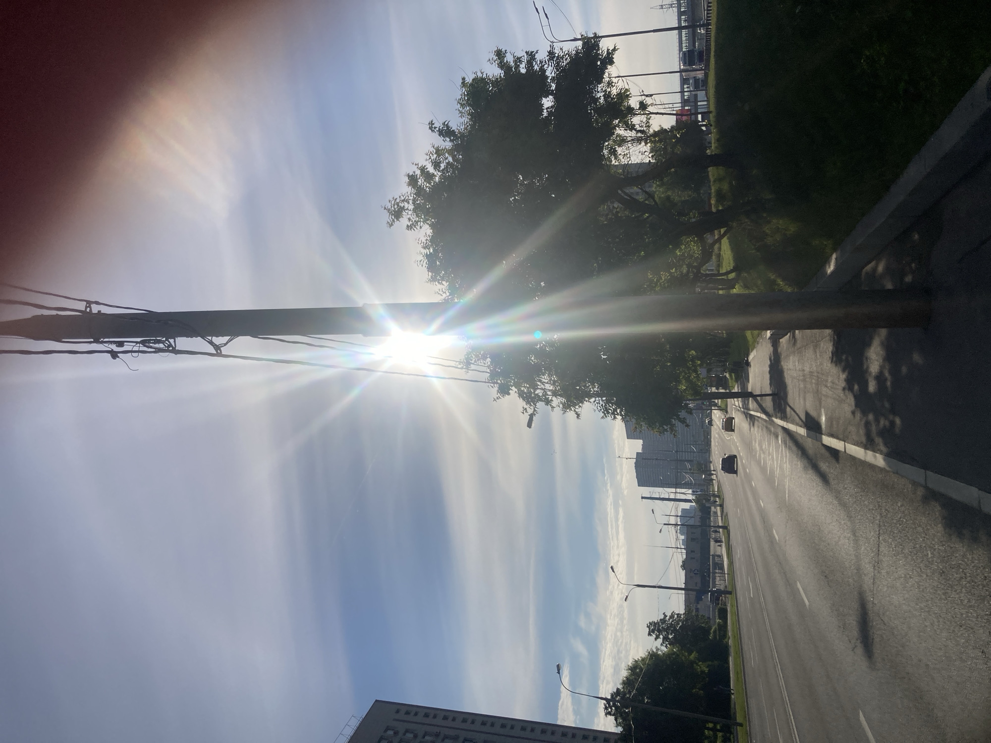 The image captures a wide, multi-lane road in what appears to be an urban or suburban setting, viewed from a slightly elevated position or sidewalk. The road extends into the distance, with a few vehicles sparsely visible, suggesting light traffic.
In the foreground, a tall utility pole or lamppost stands prominently on the left, with numerous wires and equipment attached. To the right of the pole, a large tree with dense foliage is visible. The sun is bright and low in the sky, positioned behind the utility pole and tree, creating significant lens flare and long, sharp shadows that stretch across the road. This indicates either early morning or late afternoon.
The sky is largely clear with some streaky, wispy clouds. Along the right side of the road, there is a grassy verge or median strip with additional trees and foliage. Further down the road, more trees line both sides, and various urban buildings, including a large, blocky structure on the left (when upright) and other high-rise buildings in the distance, are visible against the horizon. A large power line structure with pylons can also be seen in the far background.
No people are visible in the scene. The overall impression is one of a bright, sunny day on a major thoroughfare in a city, consistent with scenes found in Moscow.