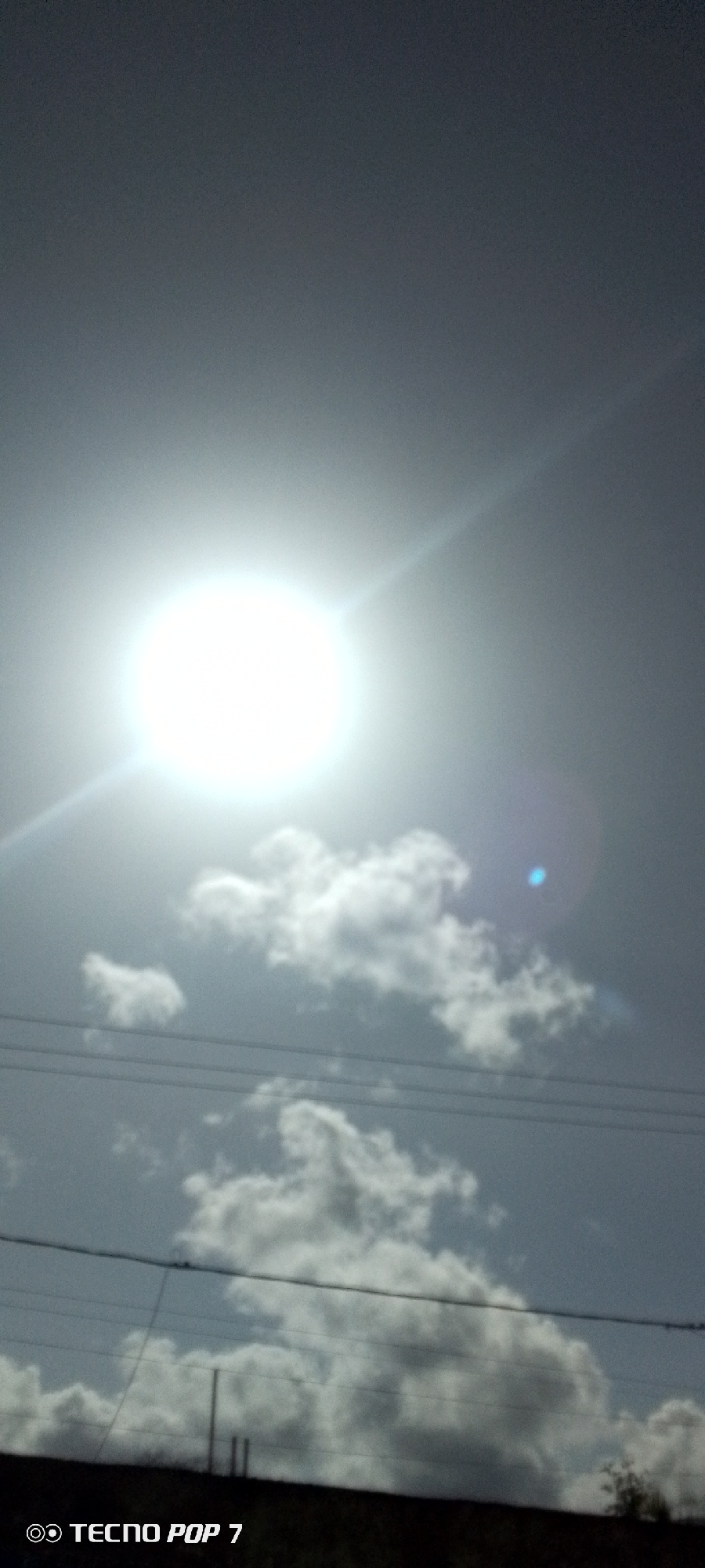 The image shows a bright sun in the sky, partially obscured by clouds. There are power lines visible in the lower portion of the frame. A blurry, dark area is at the bottom, possibly vegetation or a building. The text "TECNO POP 7" is visible in the lower left corner. The photograph has a noticeable lens flare. It is daytime. The location is specified as Altagracia, Venezuela.