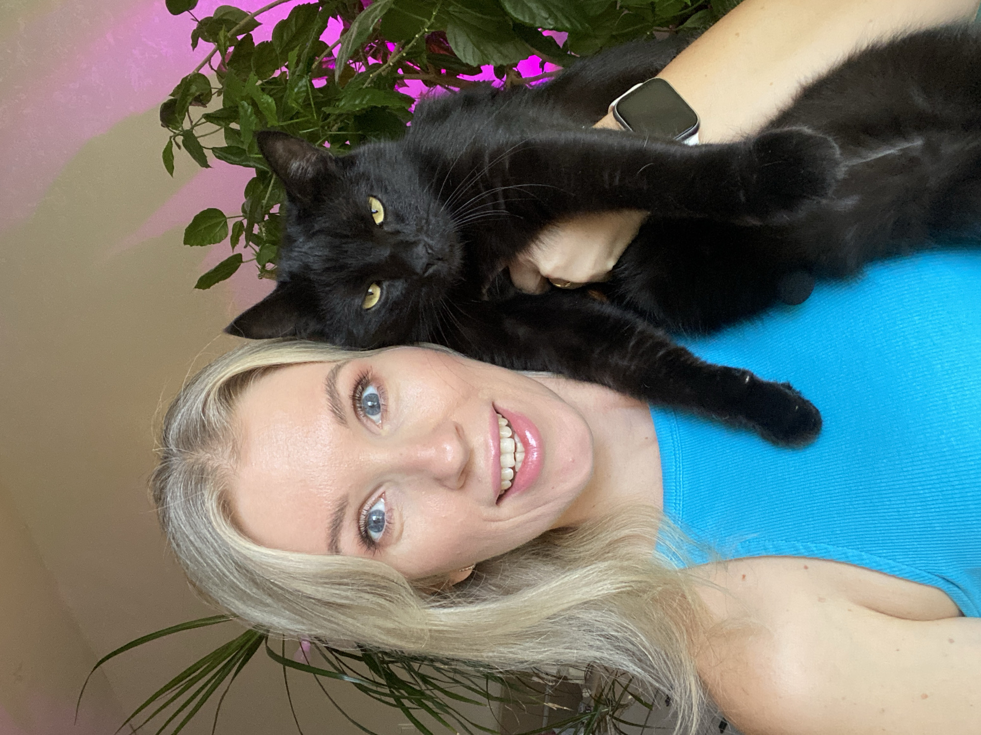 A medium shot captures a blonde woman holding a black cat in an indoor setting. The woman is positioned on the left side of the frame, facing the camera with a wide smile and bright blue eyes. She has blonde hair that falls past her shoulders. She is wearing a blue tank top. The cat, which is black with yellow eyes, is held in her right arm and faces the camera, looking slightly to its left. Behind the woman and cat, there are green plants, with a pink light illuminating the leaves of one plant on the right side of the image. A portion of a smart watch is visible on the woman's left wrist. The background appears to be a wall.