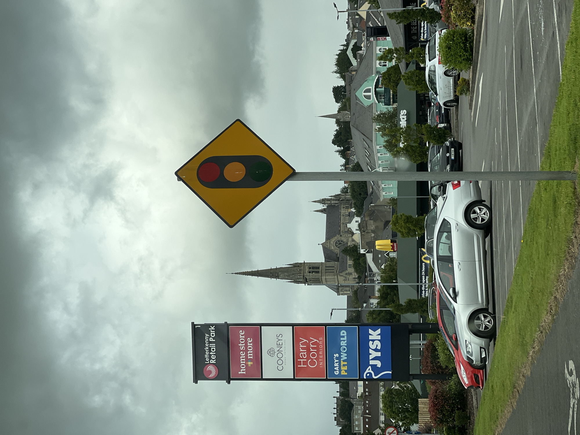 A traffic signal sign with red, yellow, and green lights is displayed on a pole in the foreground. Behind it, a large retail park sign lists several stores including Home Store + More, Cooneys Home Interiors, Harry Corry Interiors, Gary's Petworld, and Jysk. In the background, a large church with a tall spire dominates the skyline. Several cars are parked in a lot, and a McDonald's restaurant is visible. The sky is overcast.