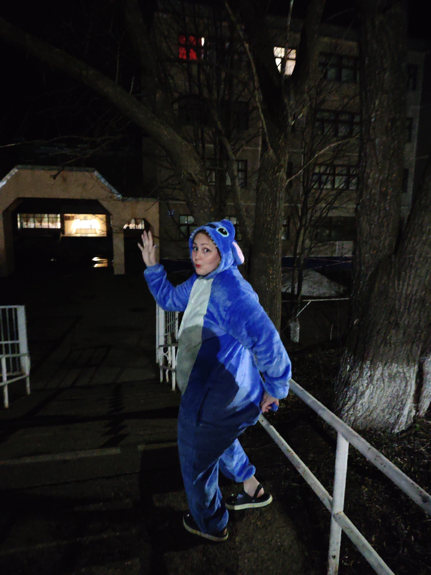 A woman in a blue "Stitch" costume stands on a set of stairs in an outdoor urban environment, likely at night or during low light conditions, in Markovskii, Russia. She is holding onto a white metal railing and waving with her right hand. The background shows a building with illuminated windows and a large tree with bare branches. A smaller structure with an open entrance is visible to the left. The area around the tree appears to have some ground cover.