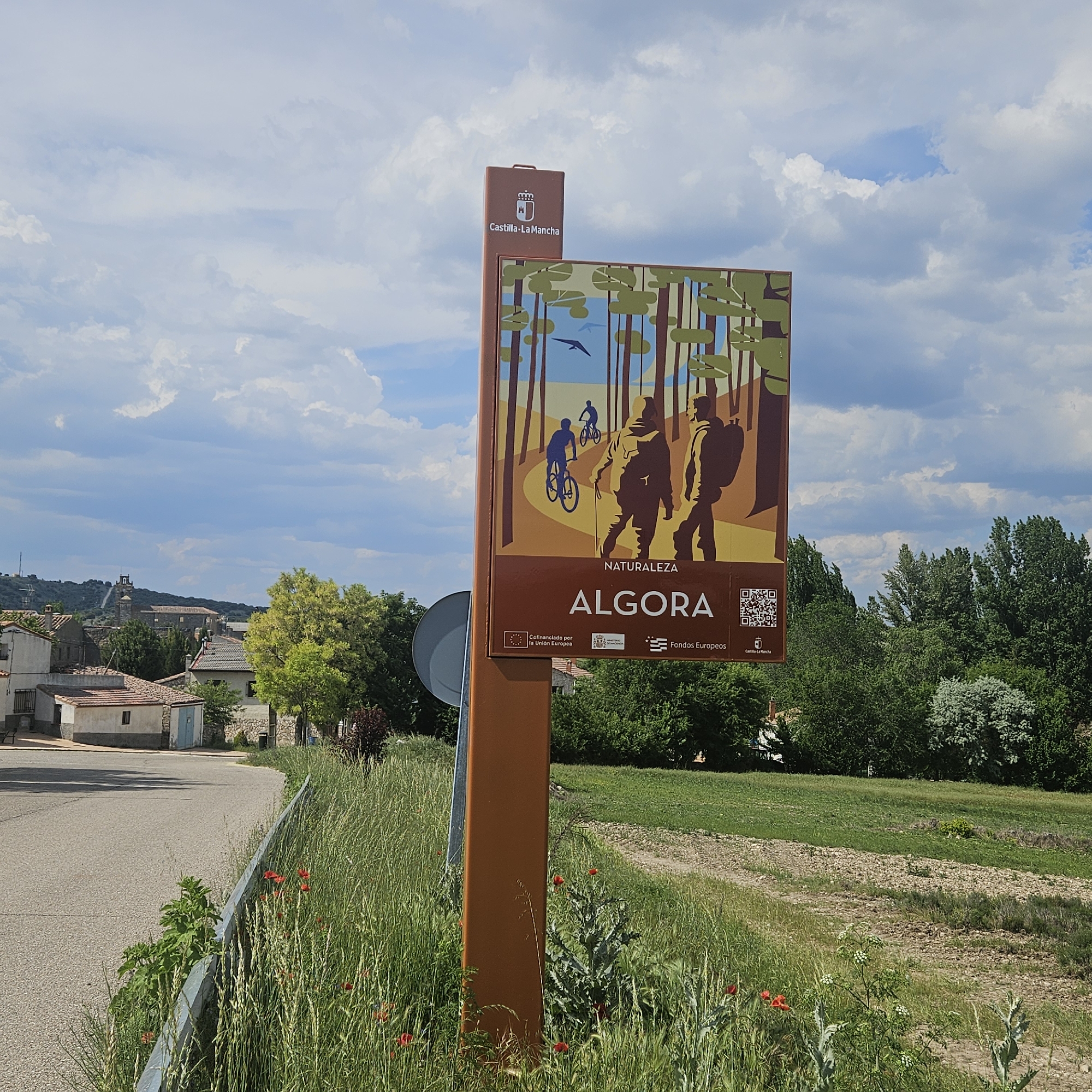 The image shows a tourism sign for the town of Algora, Spain. The sign is on a brown metal post, and the sign itself is a similar shade of brown. It features a stylized illustration of people hiking and biking through a forest, with the words "NATURALEZA ALGORA" printed in white. The Castilla-La Mancha regional logo is at the top of the post. There are also logos for the European Union and the Castilla-La Mancha government, indicating the project is co-funded by them. A QR code is also on the sign.
The sign is located next to a road, with tall grass and wildflowers growing along the roadside. In the background, there are houses and trees, suggesting the sign is located in a rural or suburban area of the town. The sky is cloudy with occasional patches of blue.