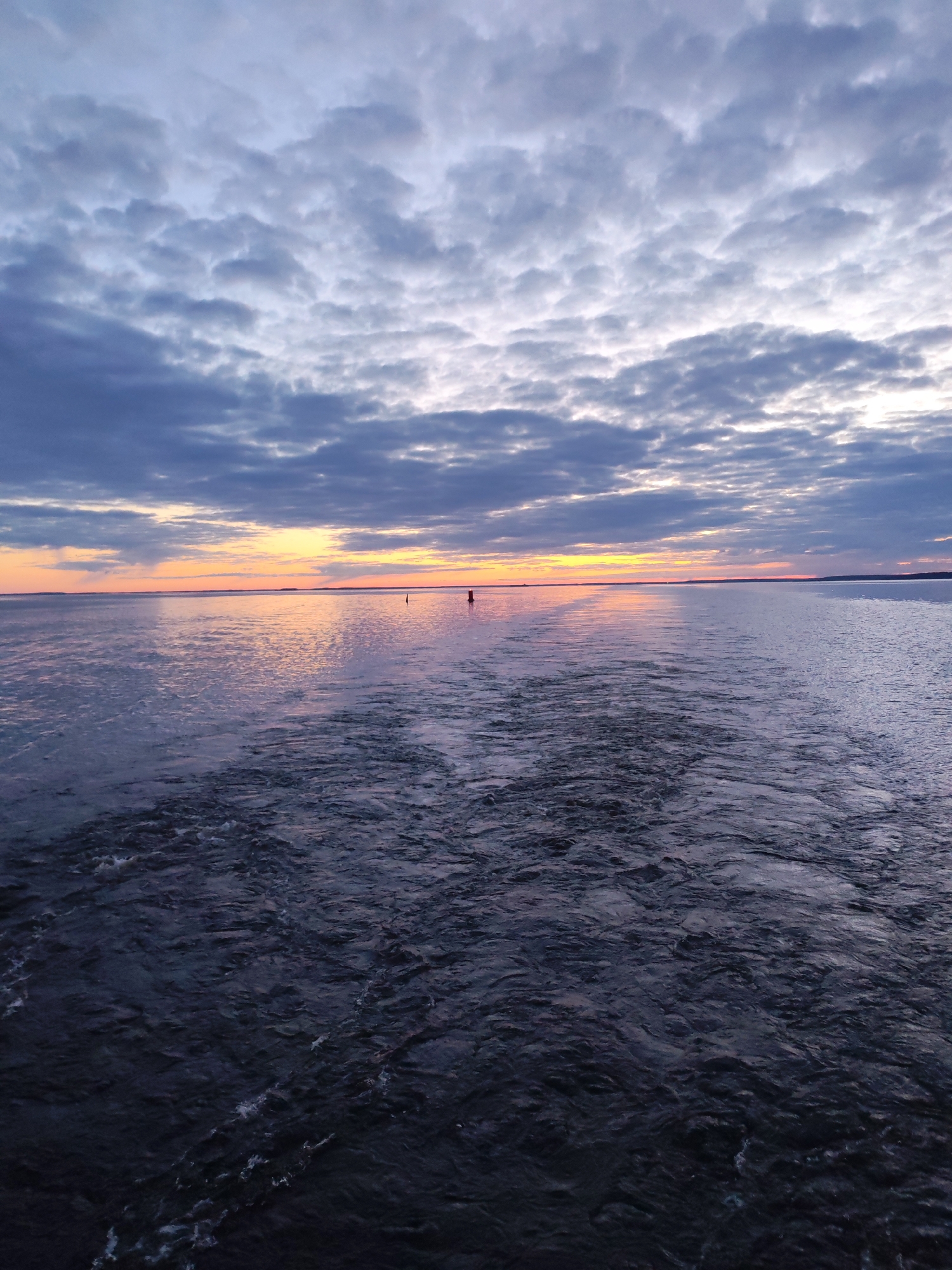 The image depicts a body of water under a cloudy sky at sunset. A wake indicates movement across the water's surface. A red navigational buoy is visible in the distance. The location is Gorushka, Russia.