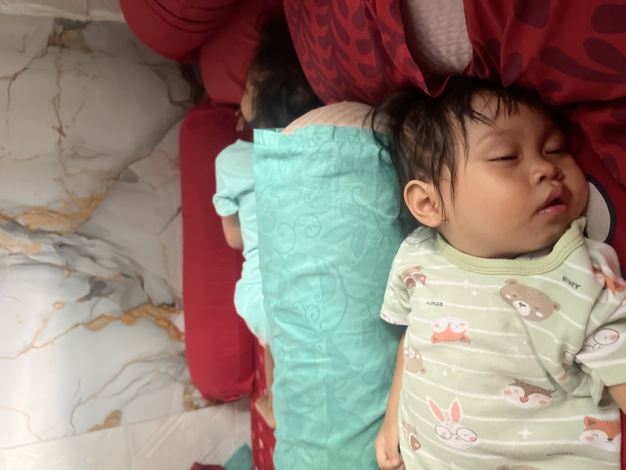 Three children are sleeping on a bed with red and green patterned sheets in a room. The child in the foreground, wearing a light green pajama set with animal prints, is positioned on their side. Behind this child, two other children are also asleep. One is wearing a turquoise sleepsuit and is partially obscured by a red bolster pillow. The third child is visible in the background, facing away from the camera. The room's wall has a marble-like pattern in white and gold. The overall scene depicts a peaceful moment of rest. The location is not discernible from the image.