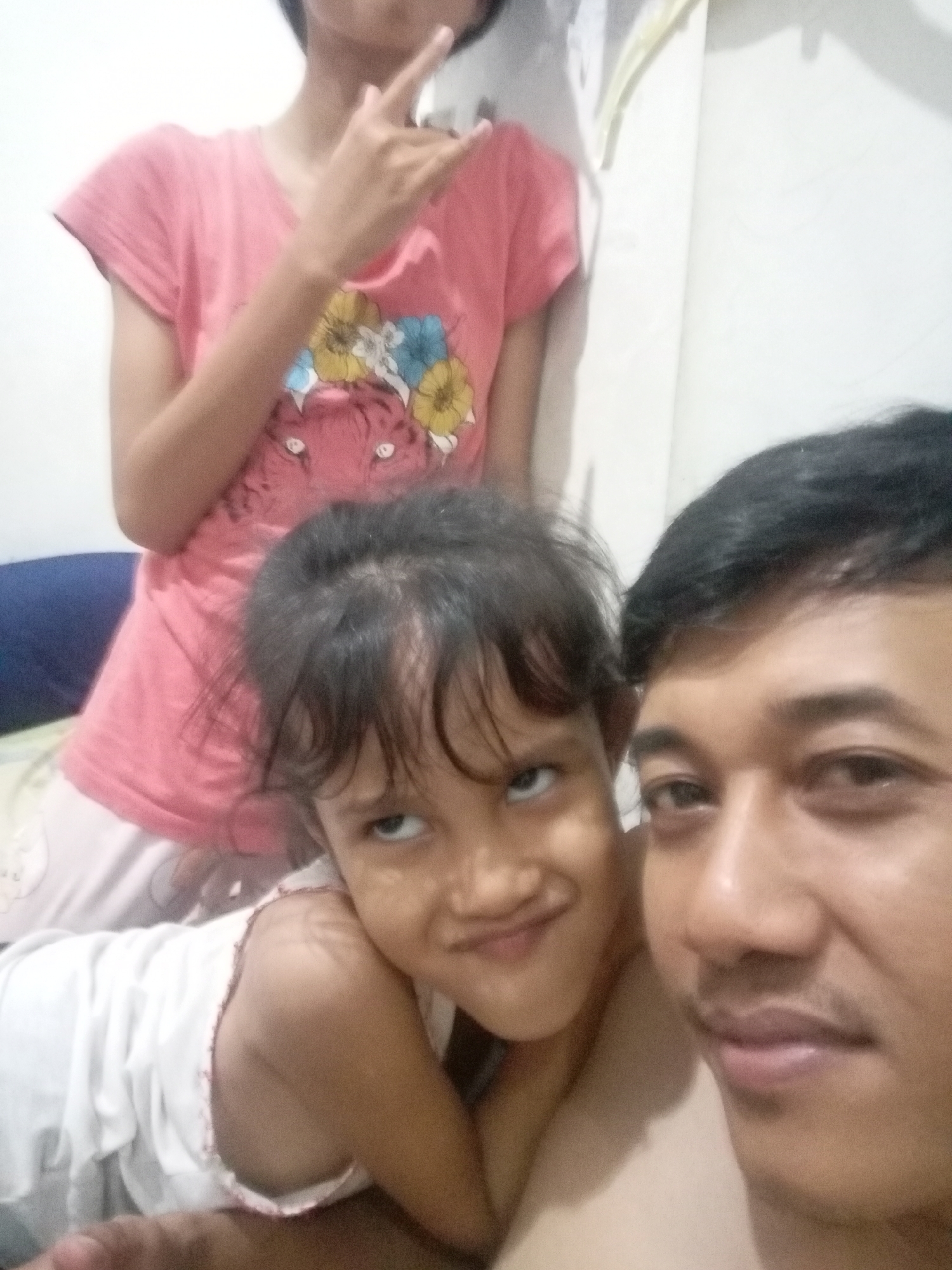 In Batam, Indonesia, a close-up selfie captures three individuals indoors. In the foreground, a man's face is partially visible on the right, with his eyes open and looking forward, and his lips slightly parted. To his left, a young girl with dark hair is looking upwards and to her left, with a slight smile. She is wearing a white garment. Behind the girl, a third person, appearing to be a young woman or older girl, is standing and partially obscured. She is wearing a pink t-shirt with a graphic design featuring a tiger and floral elements. Her left arm is raised, with her hand in a "rock on" gesture. The background consists of a plain wall and a hint of a blue surface, likely furniture or bedding. The lighting suggests an indoor setting, possibly a bedroom.