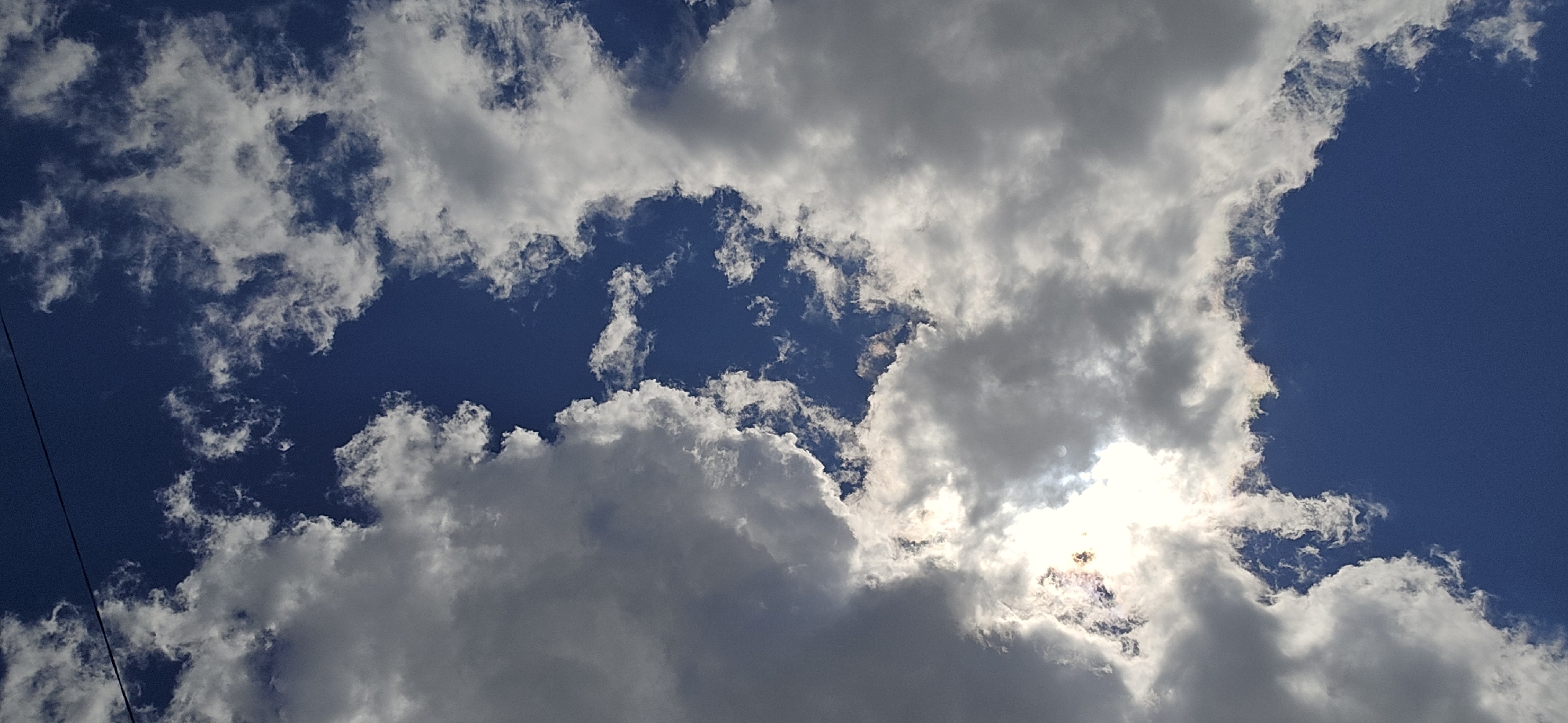 The image is a vertical shot of a partially cloudy sky. The dominant colors are the deep blue of the sky and the white and gray of the clouds. The sun is partially obscured by clouds in the upper right quadrant, creating a bright halo effect with some iridescent colors visible. The clouds are varied in texture, with some appearing fluffy and white, while others are darker and more dense. There are no visible people, structures, or specific landmarks to indicate a location in Elbrus, Russia. The image appears to be a close-up, looking directly upwards.