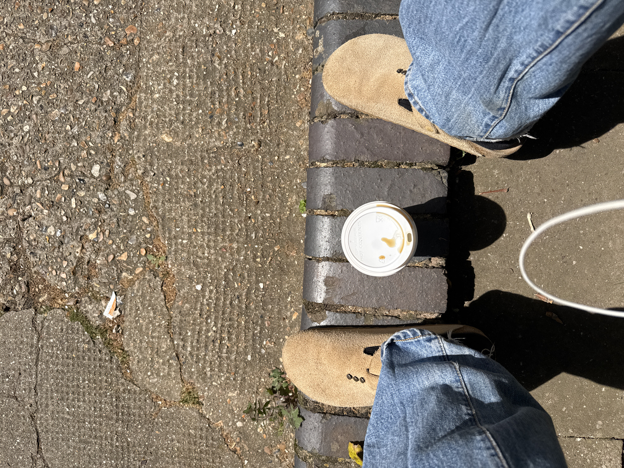 The image depicts a person standing outdoors on a city street. The person is wearing light blue jeans and tan suede shoes with visible black accents. A white, disposable coffee cup with a lid sits between the person's feet on a dark grey brick surface. The street surface is primarily cracked, textured concrete. The location is London, United Kingdom.