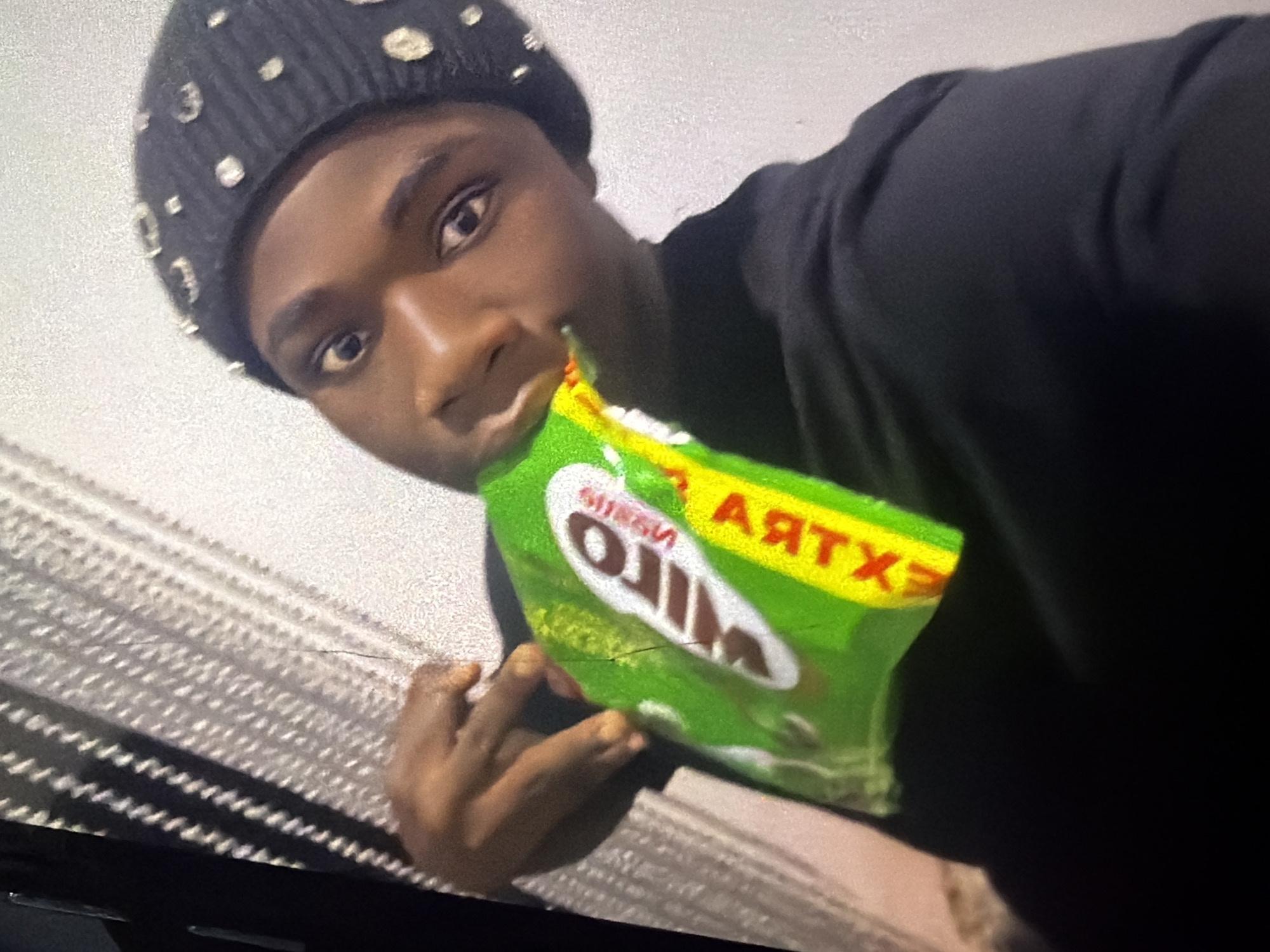 A young Black person wearing a black beanie adorned with rhinestones and a black t-shirt is captured in a selfie. They are holding a green and white bag of chips with red and yellow lettering to their mouth, appearing to be eating from it. The background is a textured white wall, and to the left, there is a dark object casting a striped shadow across a light-colored, textured surface. The image's resolution is grainy. The provided context places the scene in Bukuru, Nigeria.