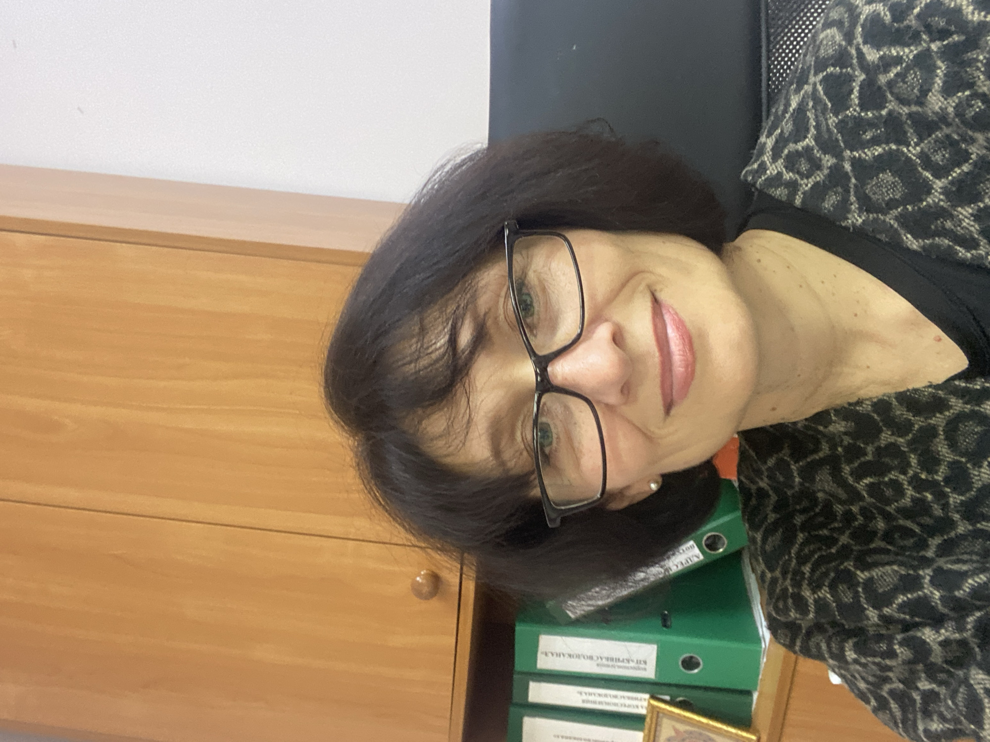 The image displays a middle-aged woman with dark, shoulder-length hair and black-framed glasses, smiling gently towards the camera. She has light-colored eyes, possibly green or hazel, and her lips are subtly colored with a pinkish hue. She wears a black top underneath a black and beige/brown leopard-print cardigan or jacket. A small, pearl-like earring is visible in her right ear.
The setting appears to be an indoor office or professional environment. To the left and slightly behind her, a light-brown wooden cabinet or desk with a visible handle is present. On the right side of the image, behind the woman, a stack of green binders is visible on the cabinet. A dark-colored office chair with a mesh back is partially seen further to the right. At the very bottom right, a small, gold-framed picture or decorative item rests near the binders. The background consists of a plain white wall on the upper left and a dark, possibly black, surface behind her head on the right.
The woman's expression is pleasant and calm, suggesting a friendly demeanor. The image is likely a casual portrait or selfie taken in her workplace.
Visible Cyrillic text on the green binders provides context. On one binder, partially obscured text reads "...НА. РАДОВАЛЬНА". Another binder clearly displays "КВ-КРПІВКАС ВОЛОЖКА.А.Л.". A third, partially visible binder shows "А. КОМПЛЕКС..." and "ОБЛІК" (meaning "accounting" or "record"). Given the provided location context of Kryvyi Rih, Ukraine, "КВ-КРПІВКАС" likely refers to a local communal enterprise or organization associated with Kryvbas, the industrial region around Kryvyi Rih.