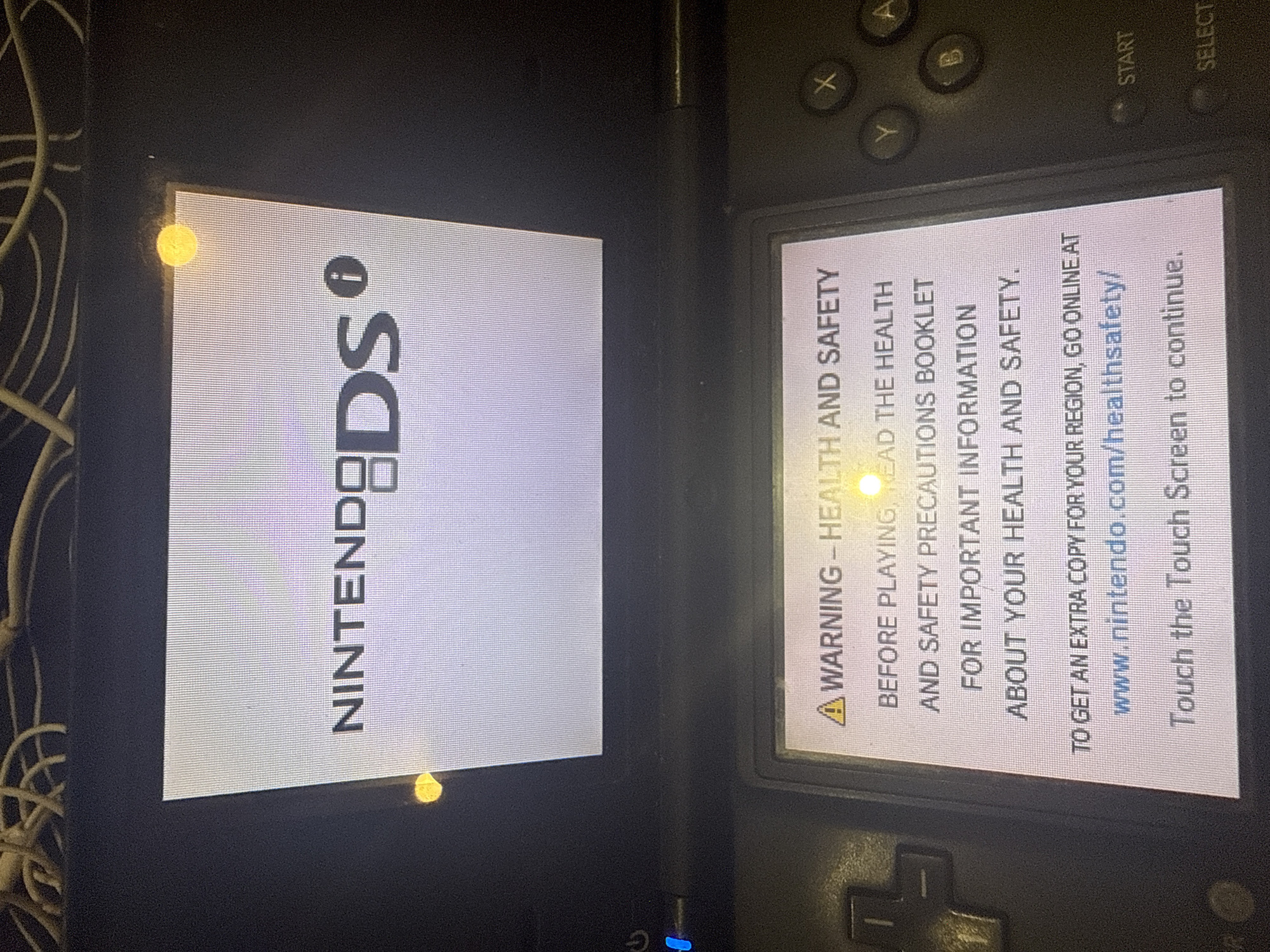 The image shows a Nintendo DS gaming console displaying its startup screen. The top screen displays the "NINTENDO DS" logo. The bottom screen shows a health and safety warning with text and a website address: "www.nintendo.com/healthsafety/". It also instructs the user to "Touch the Touch Screen to continue." The console is black and has various buttons visible. The environment appears to be indoors with some electrical wires visible in the background. There are no people or animals in the image. No information is available to determine the specific location in the city of Burgas, Bulgaria.