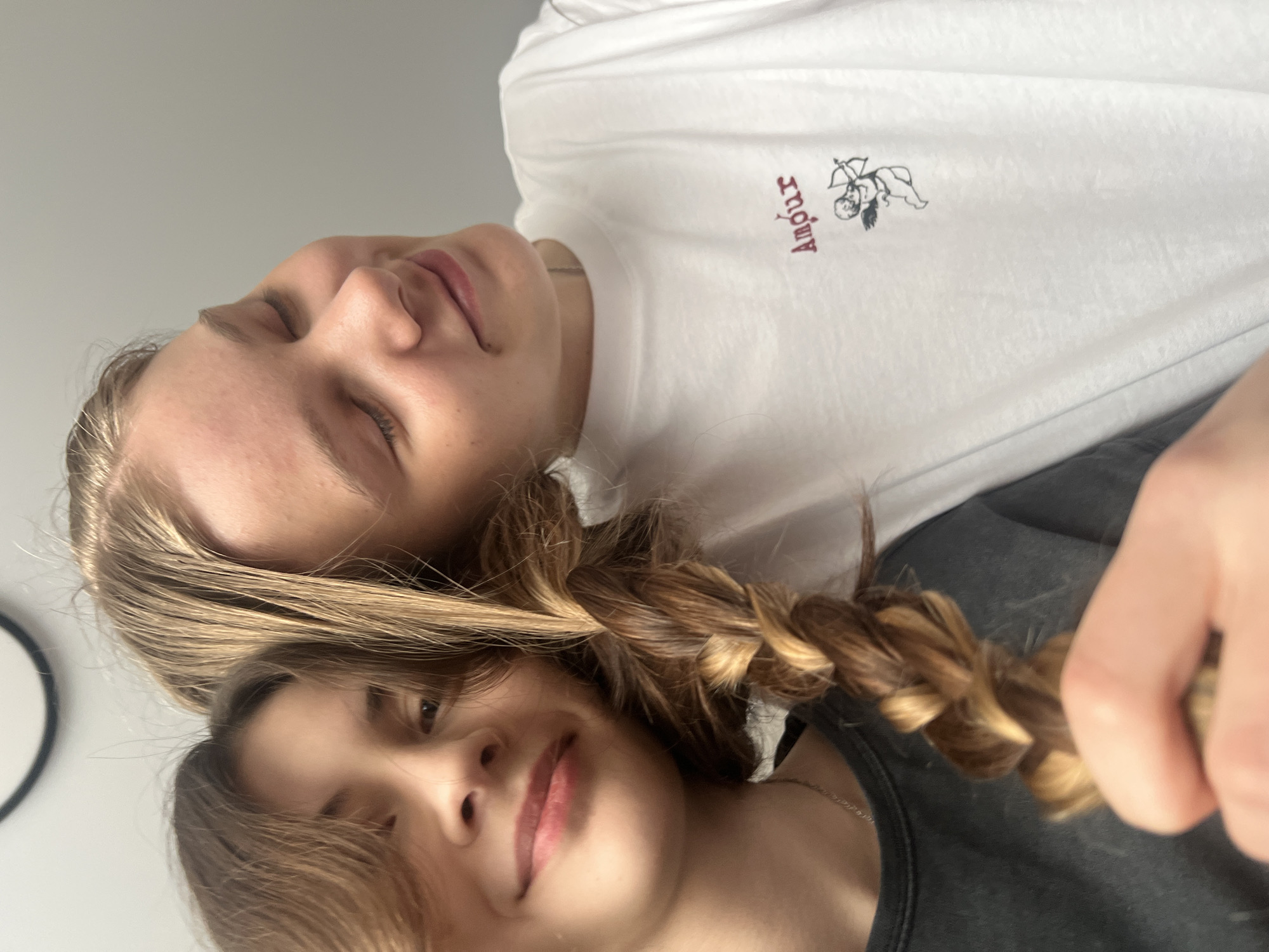 Two young individuals are closely positioned in a selfie-style photograph. The individual on the bottom has brunette hair with a braid and is wearing a black top. The individual on top has lighter hair. They are wearing a white t-shirt with "Amour" printed on it, along with an image of cupid with a bow and arrow. A circular object is visible on the left side of the image, against a plain wall. This image does not contain any information that points to a location in the city of Votkinsk, Russia.