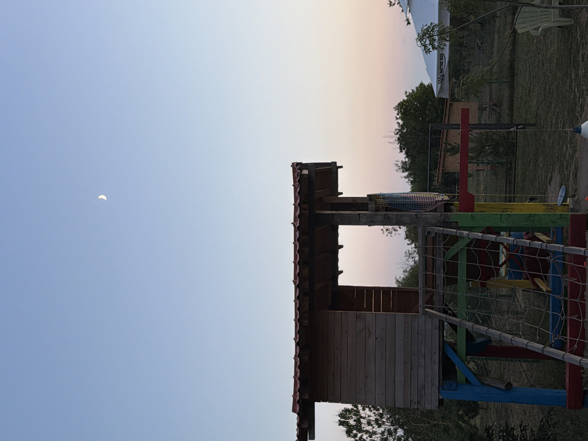 The image captures an outdoor scene at dusk in Tvarditsa, Bulgaria. The foreground features a brightly colored wooden playground structure, characterized by its red tiled roof and blue, red, green, and yellow painted supports. A rope net ladder extends from the structure. To the right, a white umbrella with Cyrillic text is visible, partially obscuring a smaller wooden building behind it. In the distance, trees are scattered across the landscape, and the sky is a gradient of pale blue and soft pink, with a crescent moon visible in the upper portion of the frame. The ground is covered in grass. The overall atmosphere is serene and twilight.