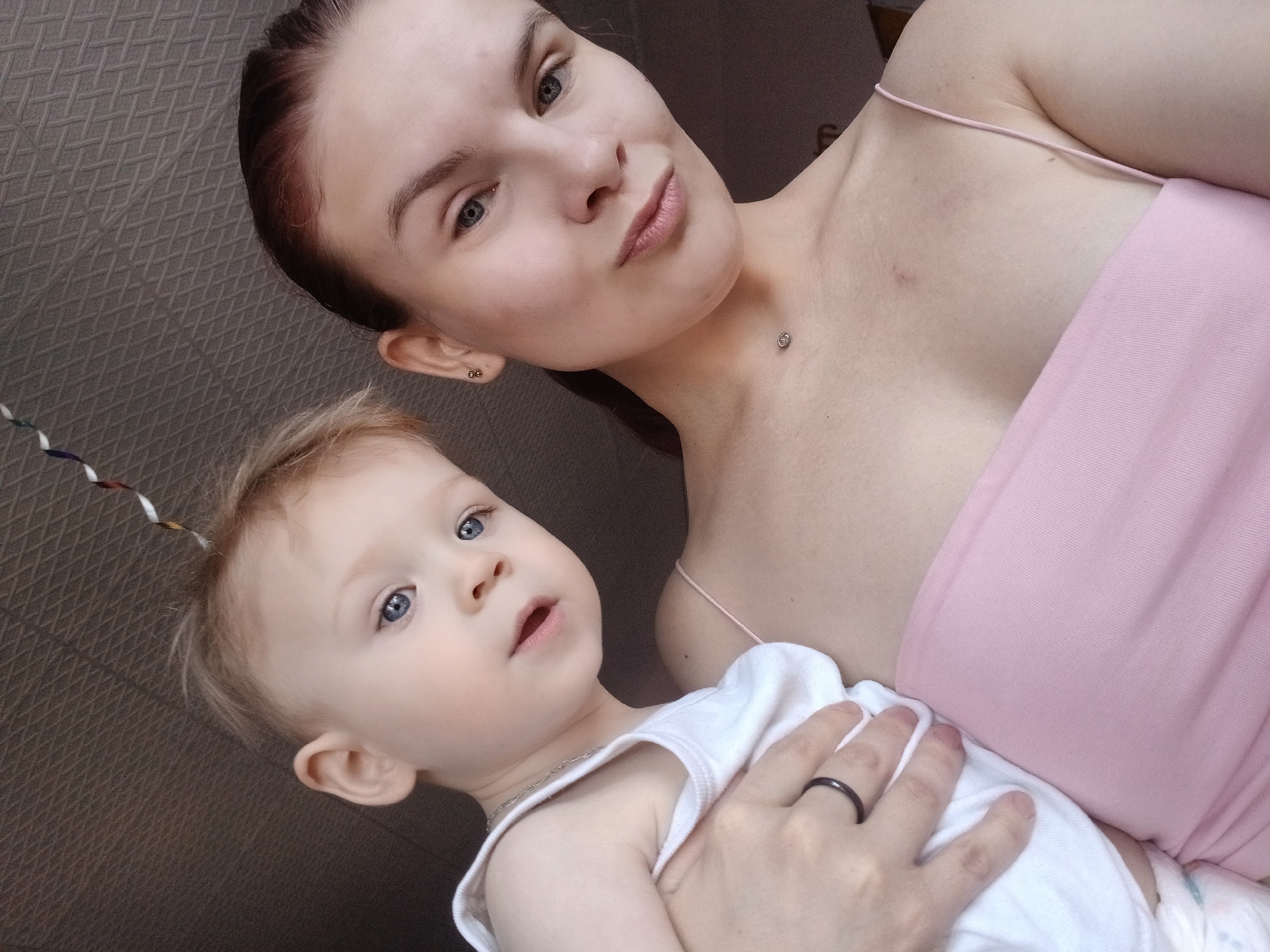 The image shows a woman and a young child in an indoor setting. The woman has fair skin, light blue eyes, and auburn hair pulled back. She is wearing a light pink top. The child has fair skin, light blue eyes, and blond hair with a colorful spiral hair accessory. The child is wearing a white tank top and is being held by the woman. The woman is wearing a dark ring on her left ring finger. The background is a textured wall. The location is presumed to be in Chaikovsky, Russia. The nature of the interaction between the woman and child seems affectionate and caring.