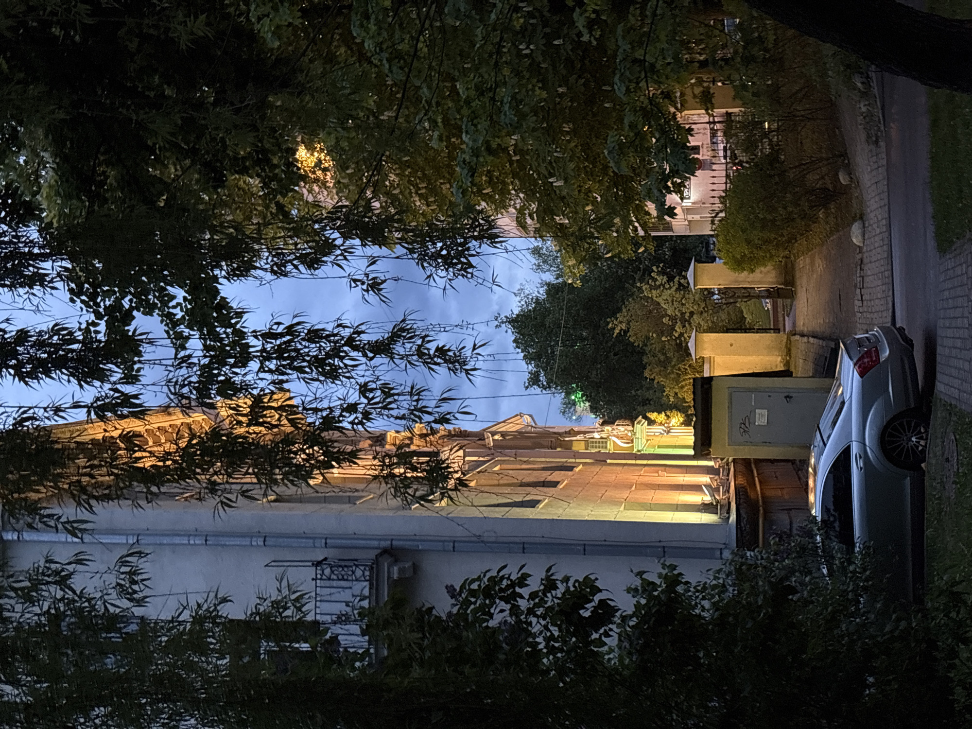 The image shows a street scene in Minsk, Belarus, at dusk. A light-colored building with multiple windows and a rooftop structure is visible through trees. A silver car is parked along the sidewalk. The sky is blue with some clouds. Streetlights illuminate the street. Green foliage is prominent in the foreground, framing the scene.