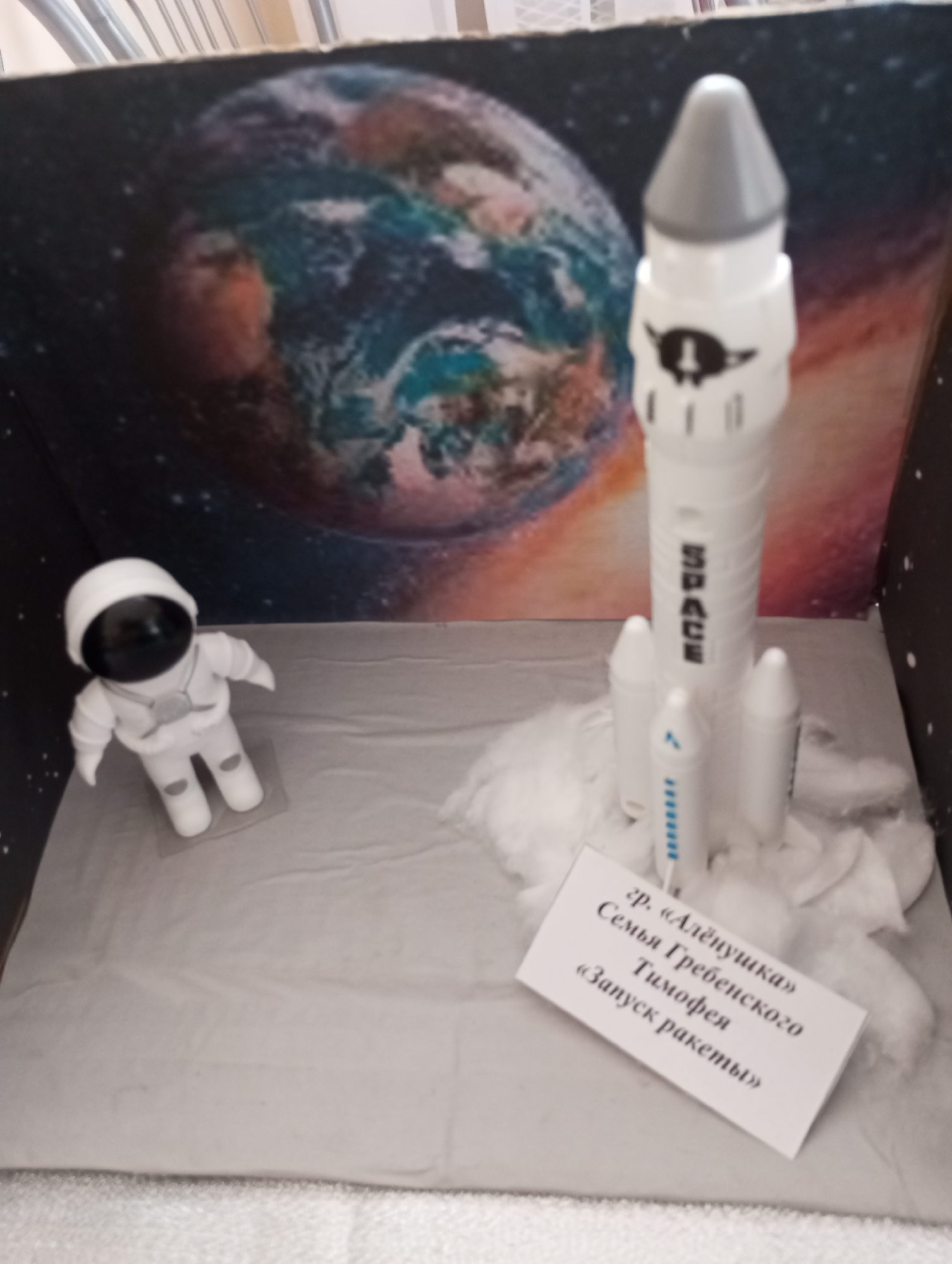 A child's diorama depicts a space launch scene. A white astronaut figurine stands to the left, and a toy rocket stands to the right surrounded by white cotton or similar material resembling exhaust. In the background, a picture of the Earth is displayed. A white card in the foreground reads (in Cyrillic script): "гр. 'Алёнушка', Семья Гребенского Тимофея, 'Запуск ракеты'", likely naming the child or family who created the diorama. The scene is contained within a box. The location of the diorama is unknown.