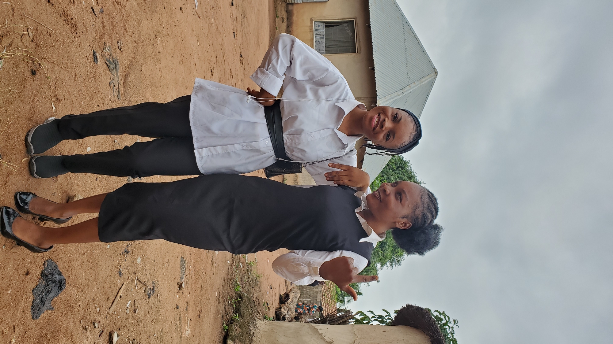 Two young women stand side-by-side in an outdoor, rural environment. The woman on the left wears a white button-down shirt with a black belt and black pants. She has her left hand on her hip and her right arm around the shoulder of the other woman. She is smiling at the camera. The woman on the right wears a white collared shirt underneath a black sleeveless dress. She has her left arm around the other woman's shoulders and her right hand is raised, with her index finger pointed upwards. She is looking to her right.
The background consists of two buildings, one with a corrugated metal roof and another with a thatched roof. The ground is covered in reddish-brown dirt, with some scattered debris. The sky is overcast. The overall setting appears to be a residential area in a village or rural community. Given the context provided about Jalingo, Nigeria, the scene likely depicts a typical residential compound in such an environment.